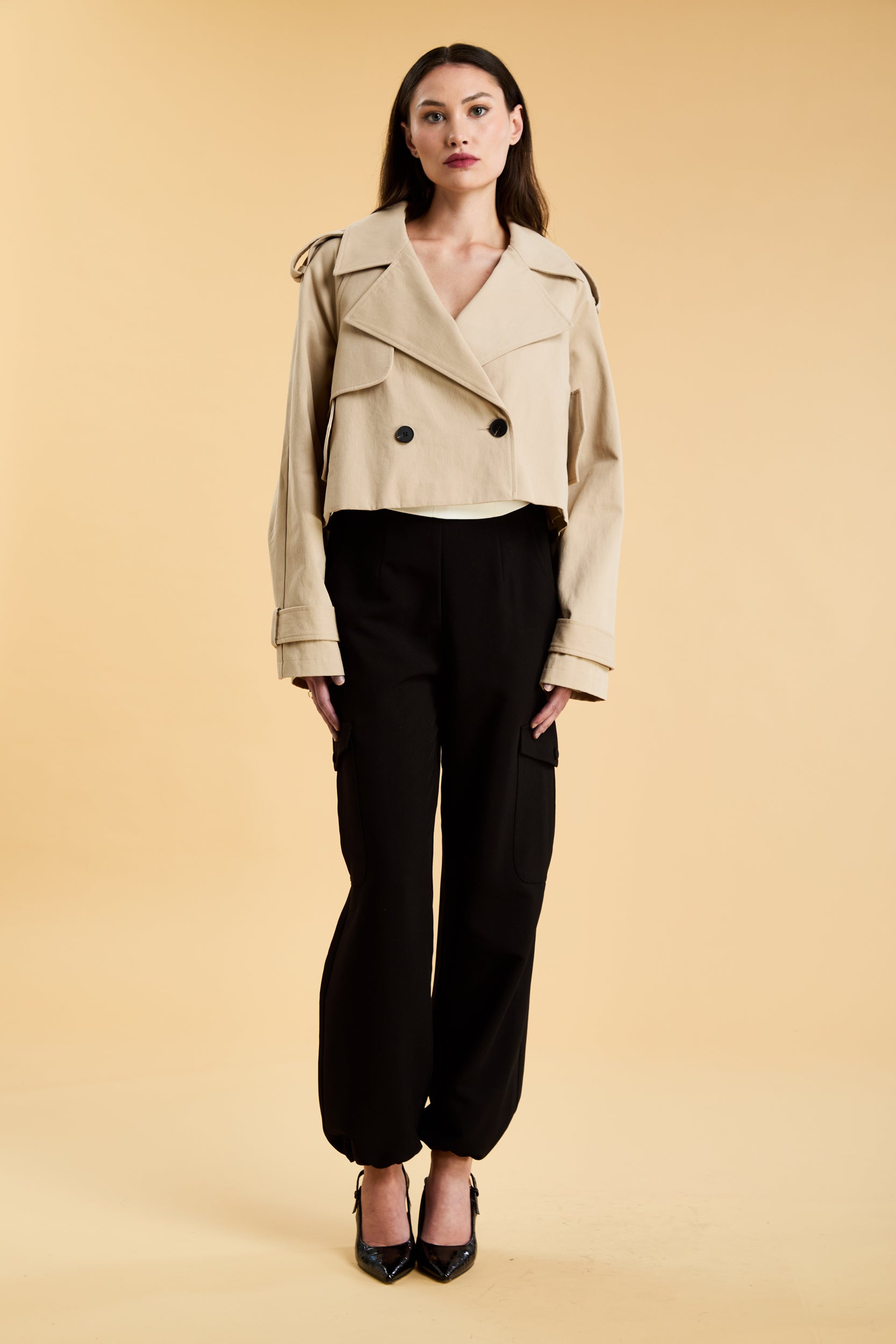 Woman wearing a beige cropped trench coat jacket in stone color against a beige background