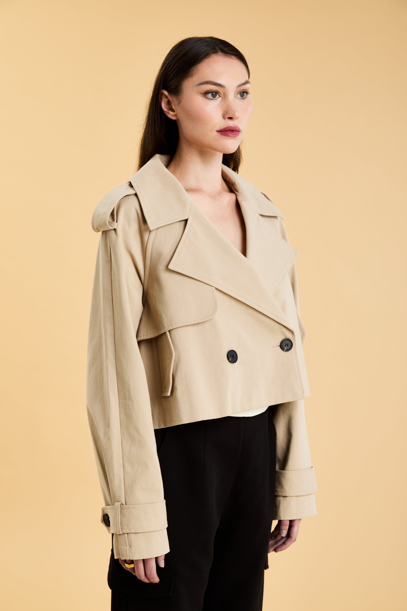 Woman wearing a beige cropped trench coat jacket in stone color against a beige background