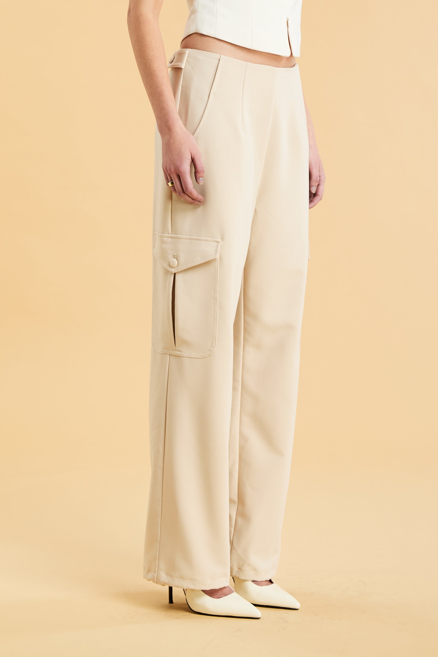 Woman wearing a white sleeveless double zipper top and beige almond Triplicity trousers cargo pants on a beige background