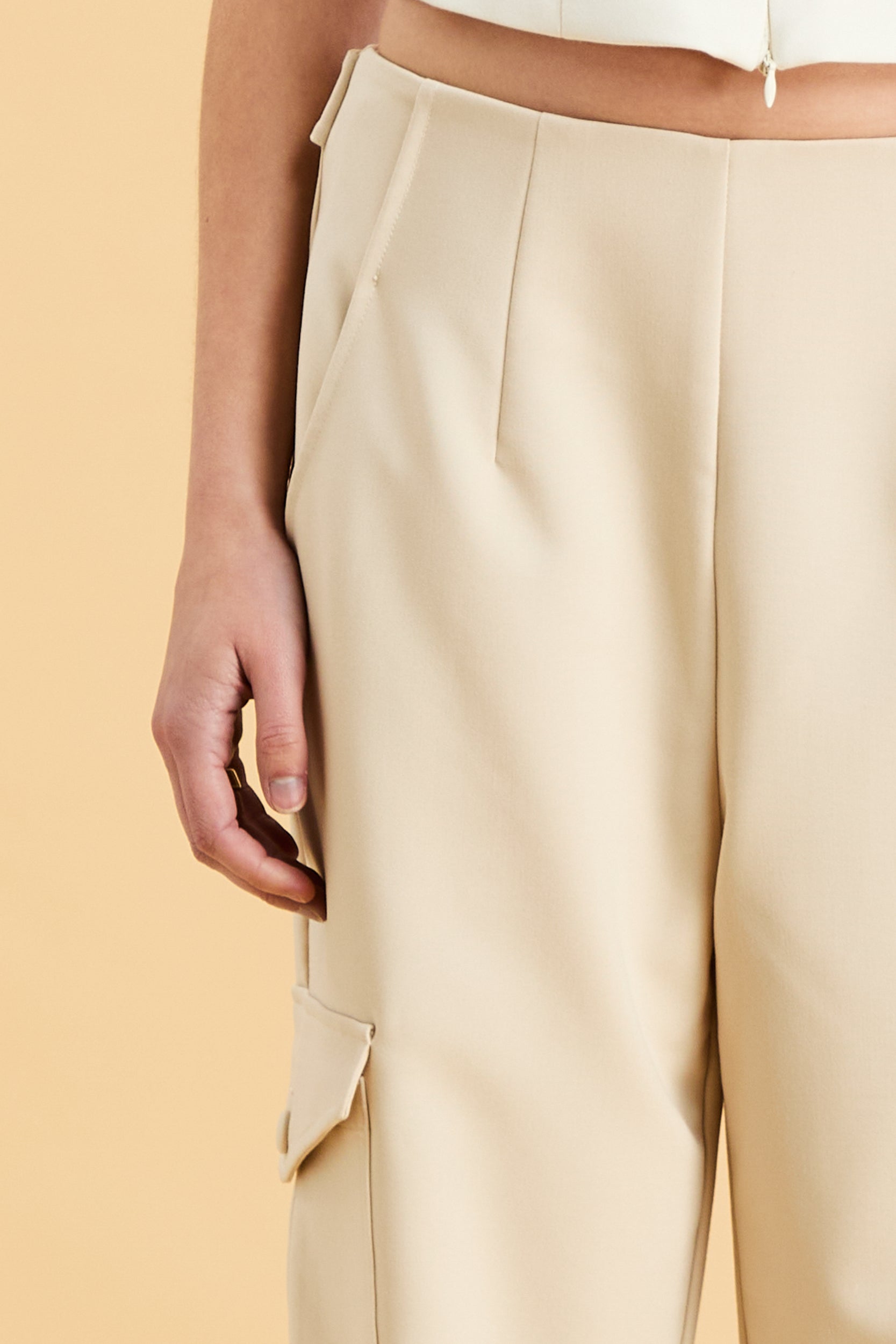 Woman wearing a white sleeveless double zipper top and beige almond Triplicity trousers cargo pants on a beige background