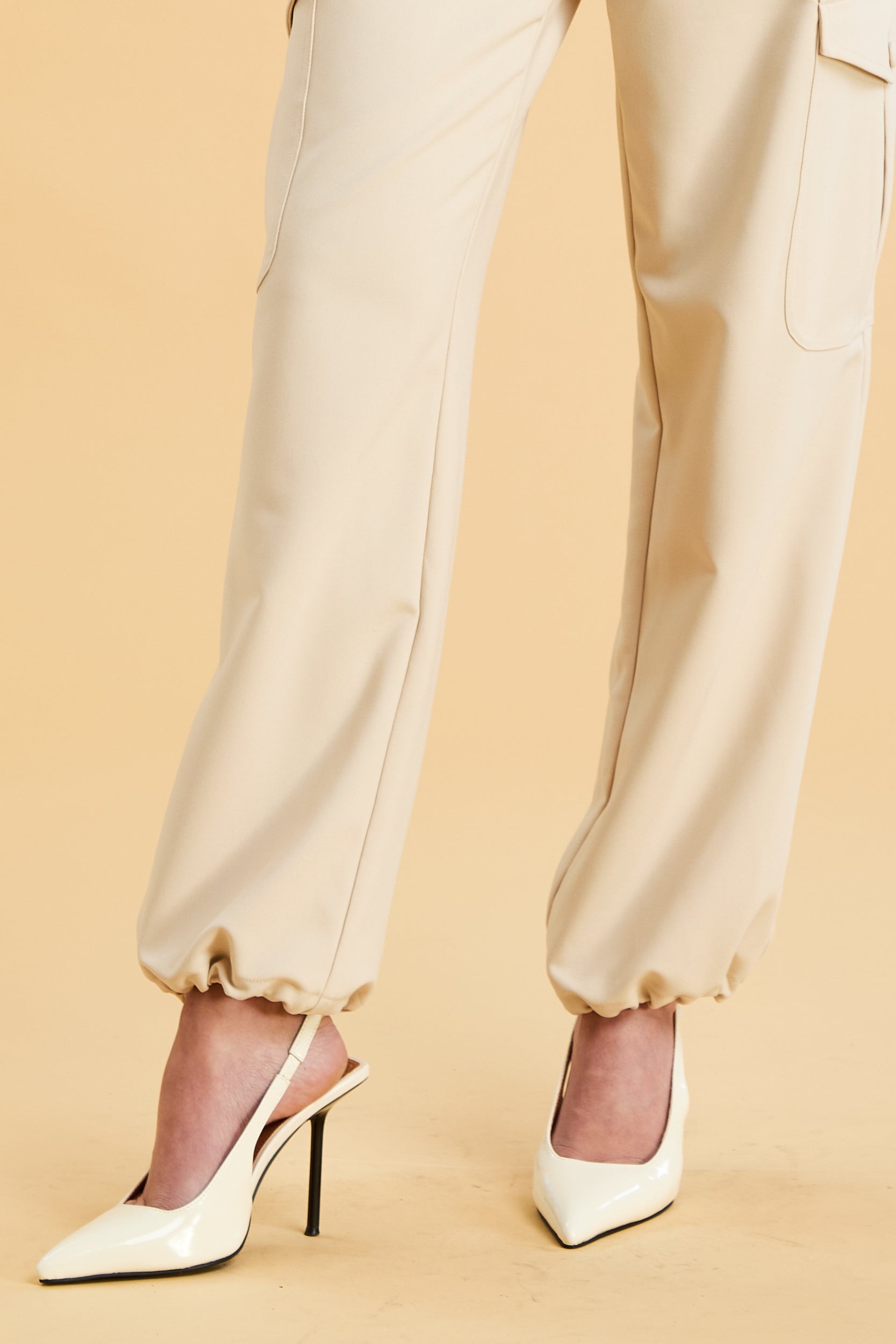 Woman wearing a white sleeveless double zipper top and beige almond Triplicity trousers cargo pants on a beige background