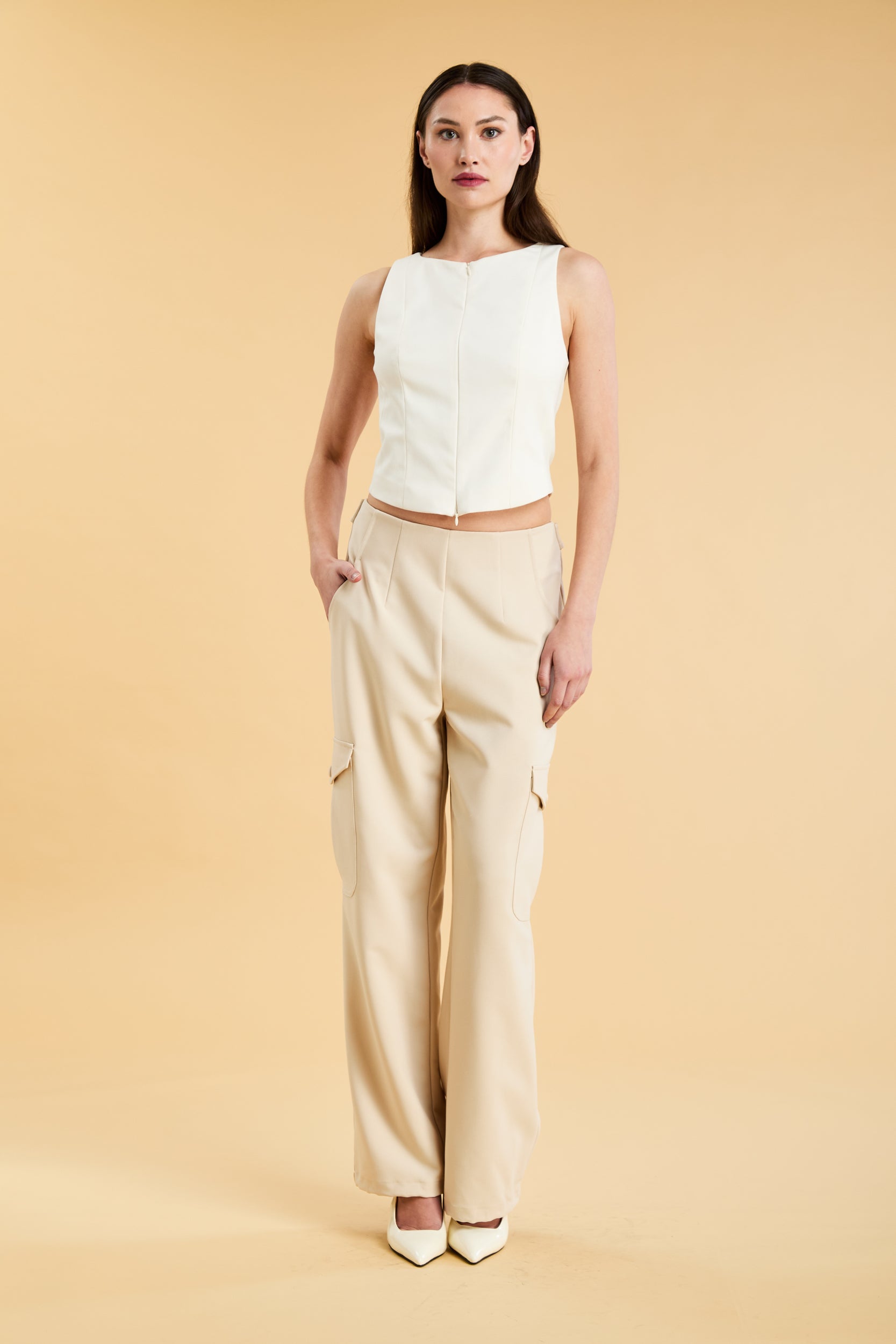 Woman wearing a white sleeveless double zipper top and beige almond Triplicity trousers cargo pants on a beige background