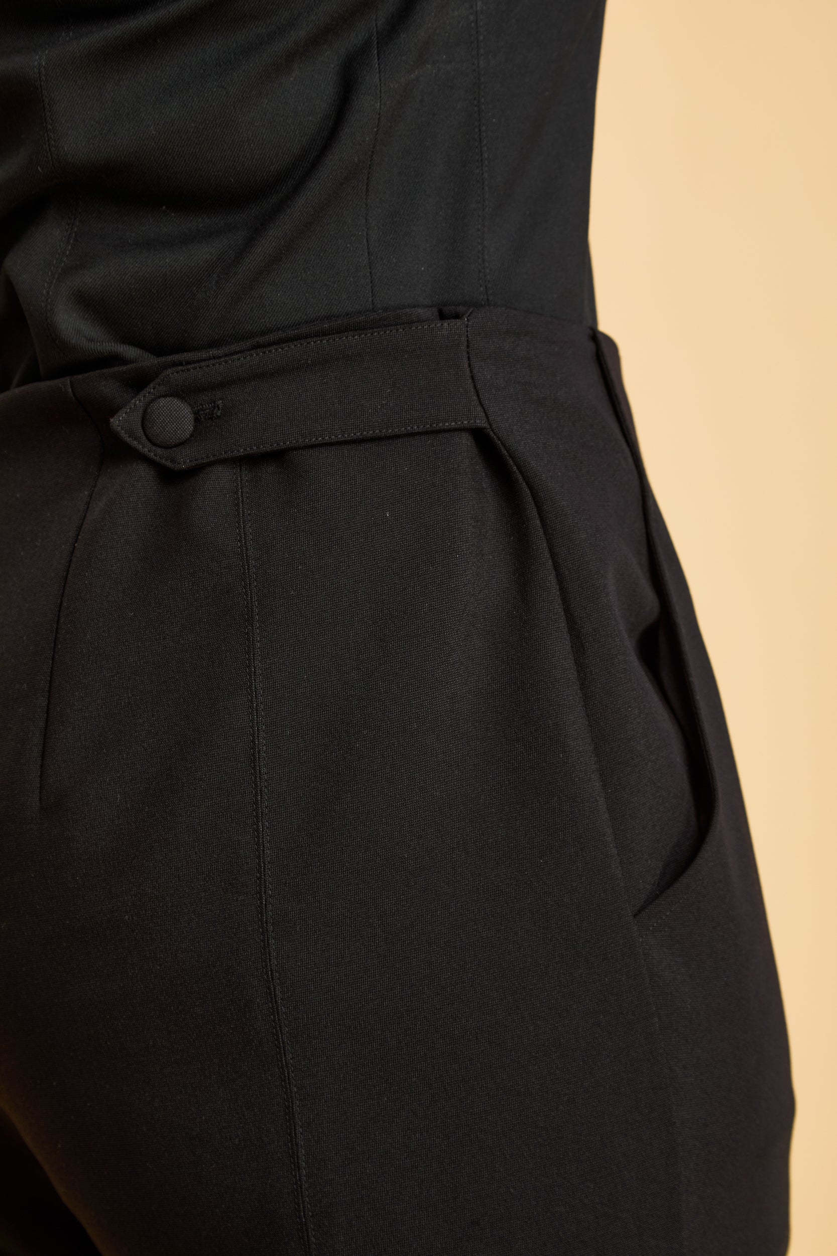 close-up of black pants with a waist tab and two buttons on a beige background