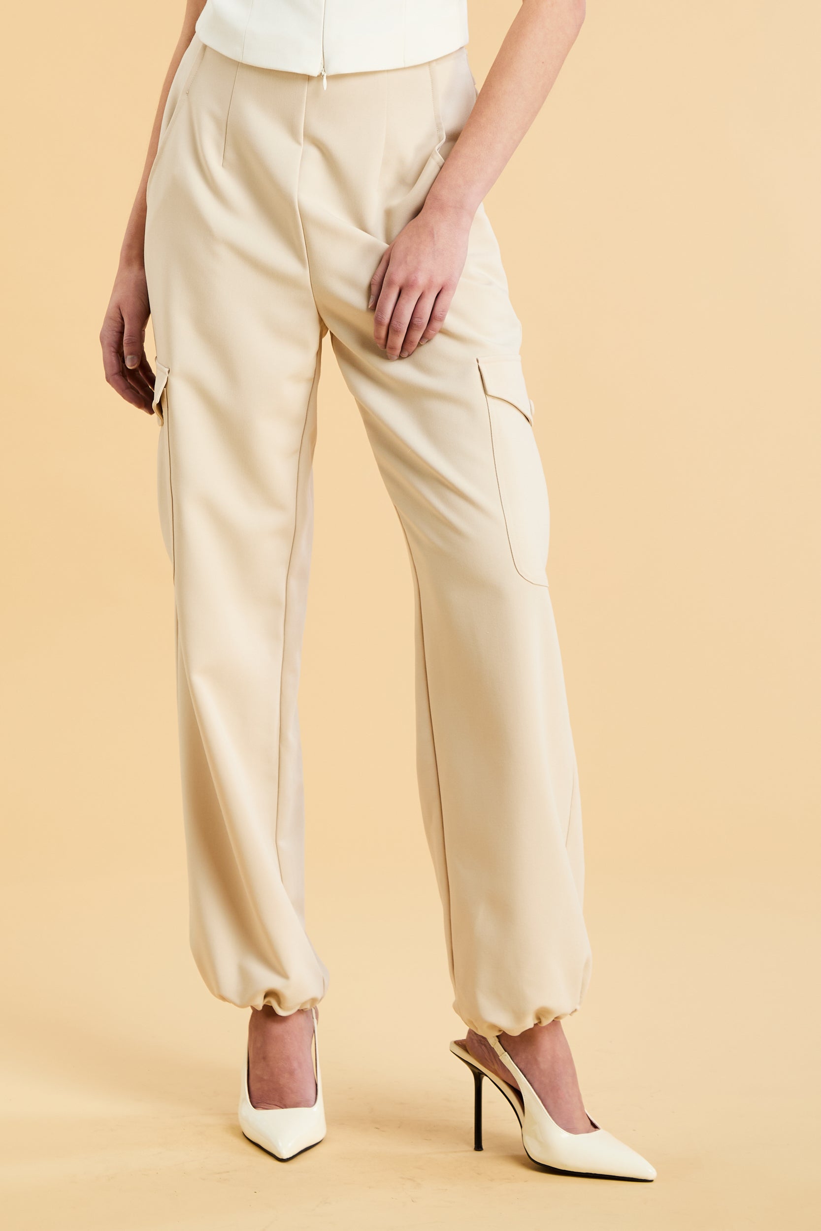 Woman wearing a white sleeveless double zipper top and beige almond Triplicity trousers cargo pants on a beige background