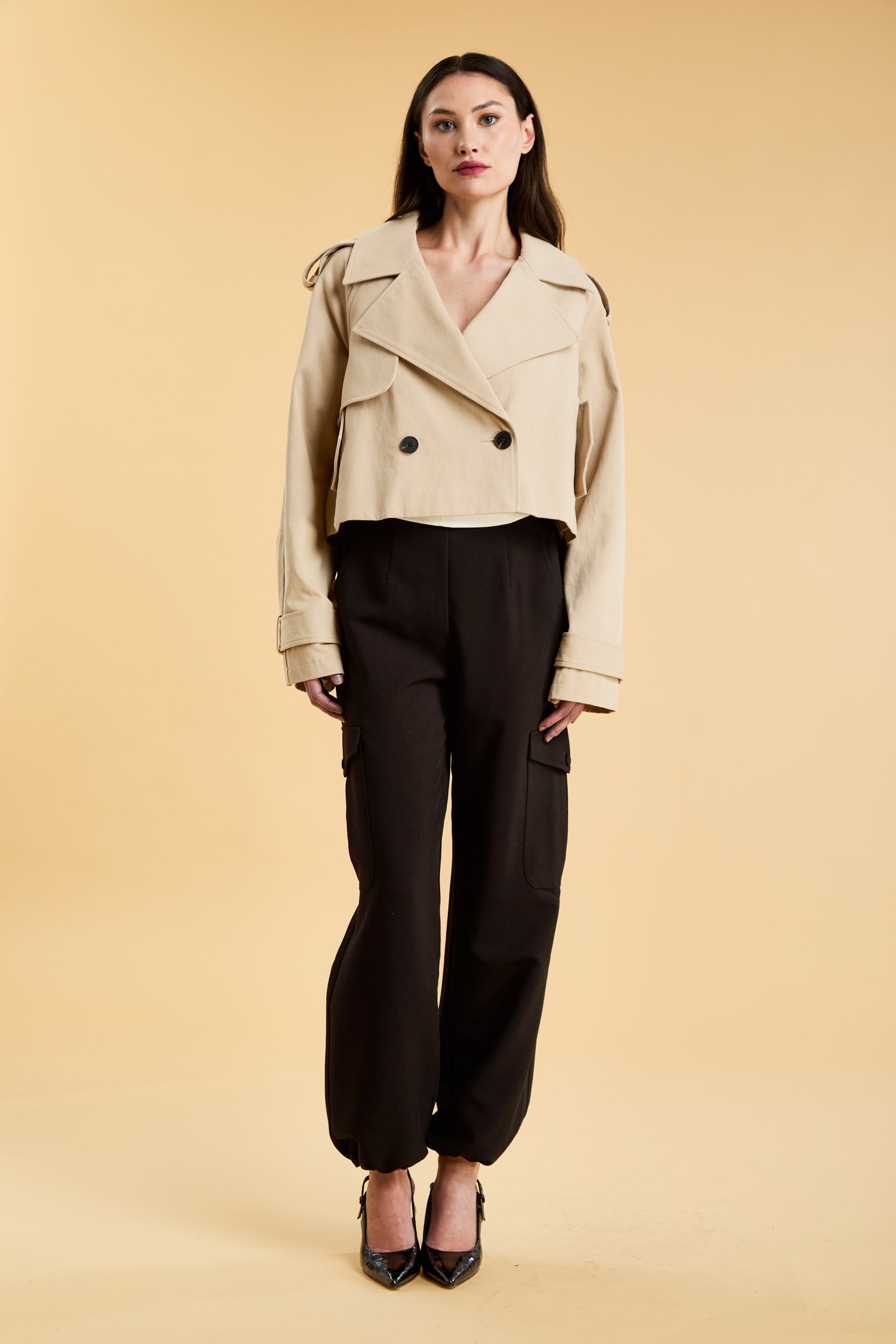 Woman wearing a cropped trench coat and black cargo pants on a beige background