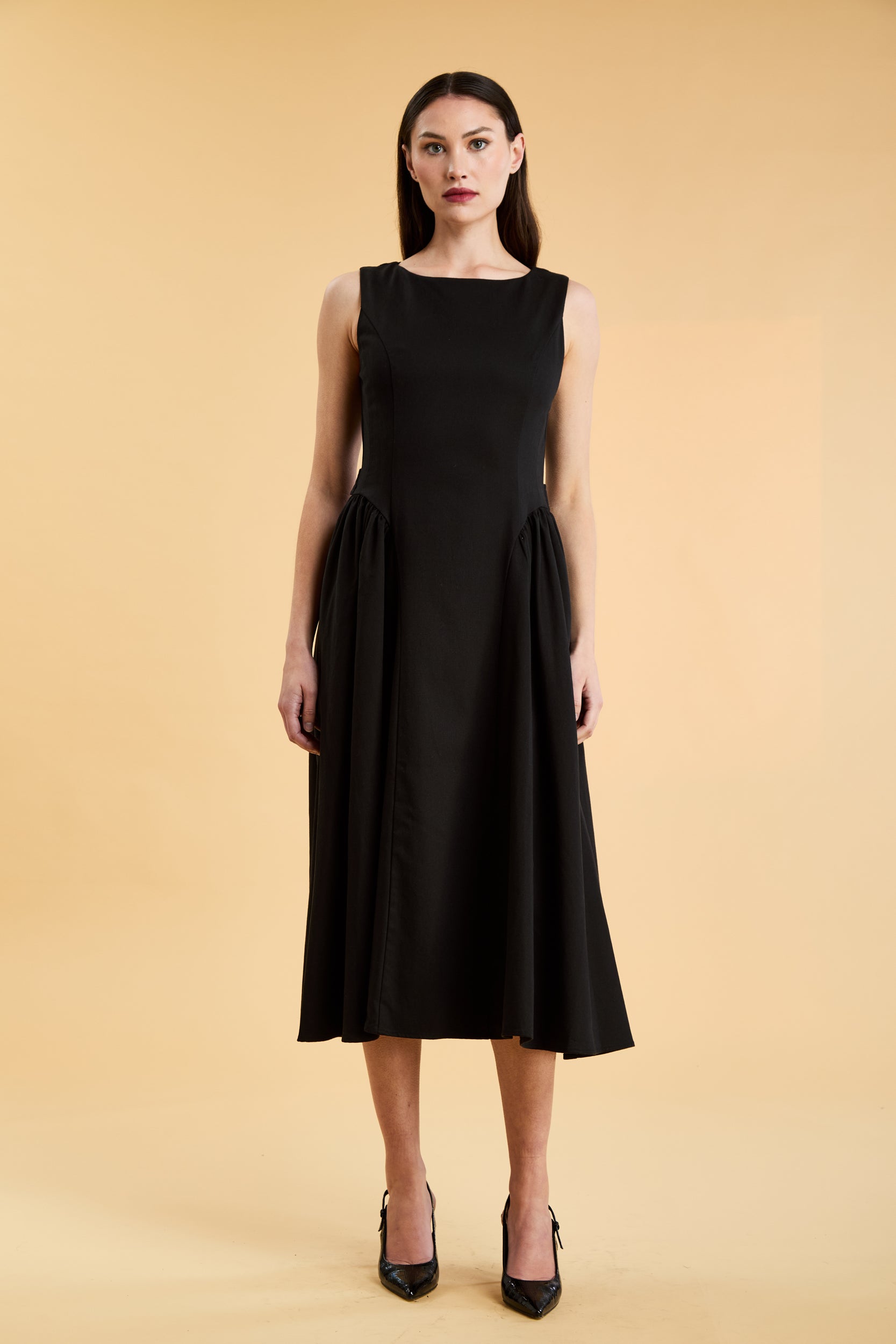 Woman wearing a Triplicity Black sleeveless midi dress with gathered side panels on a beige background