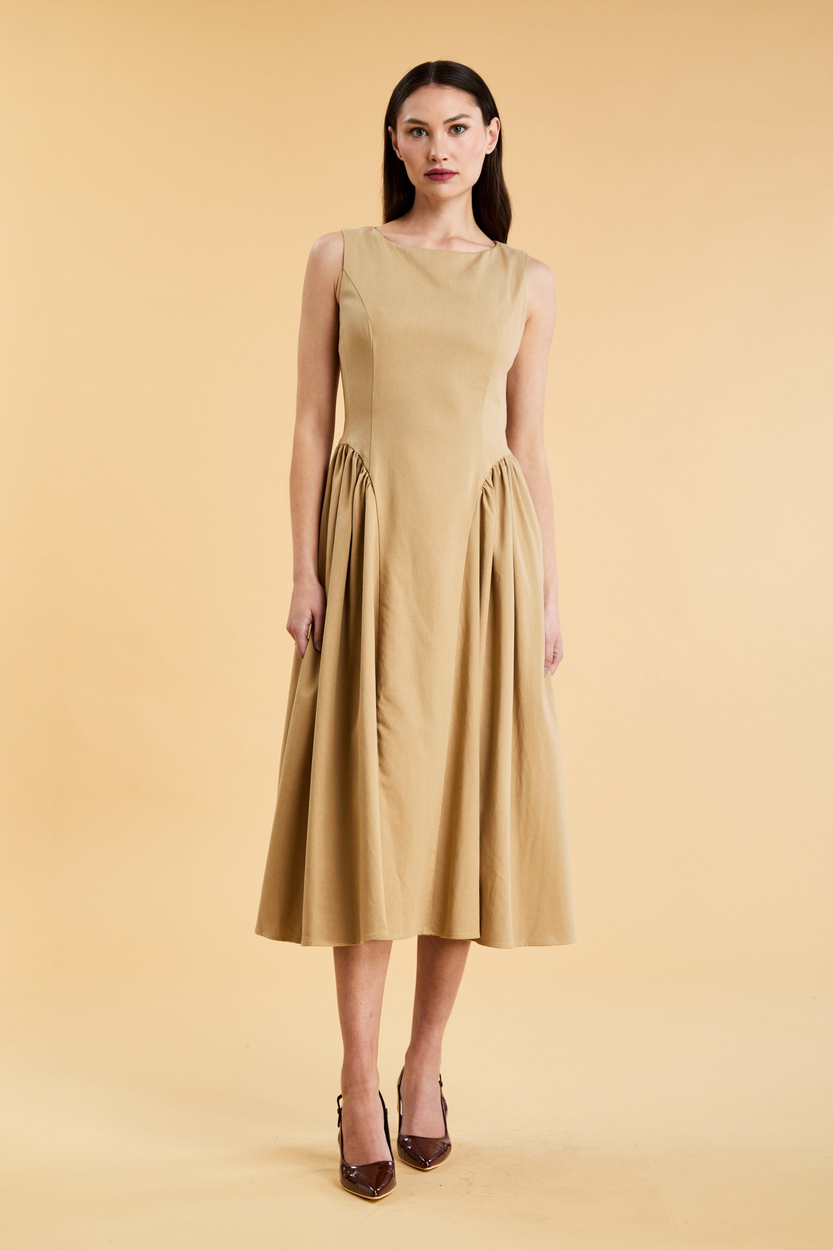 Woman wearing a Triplicity Camel colored sleeveless midi dress with gathered side panels on a beige background