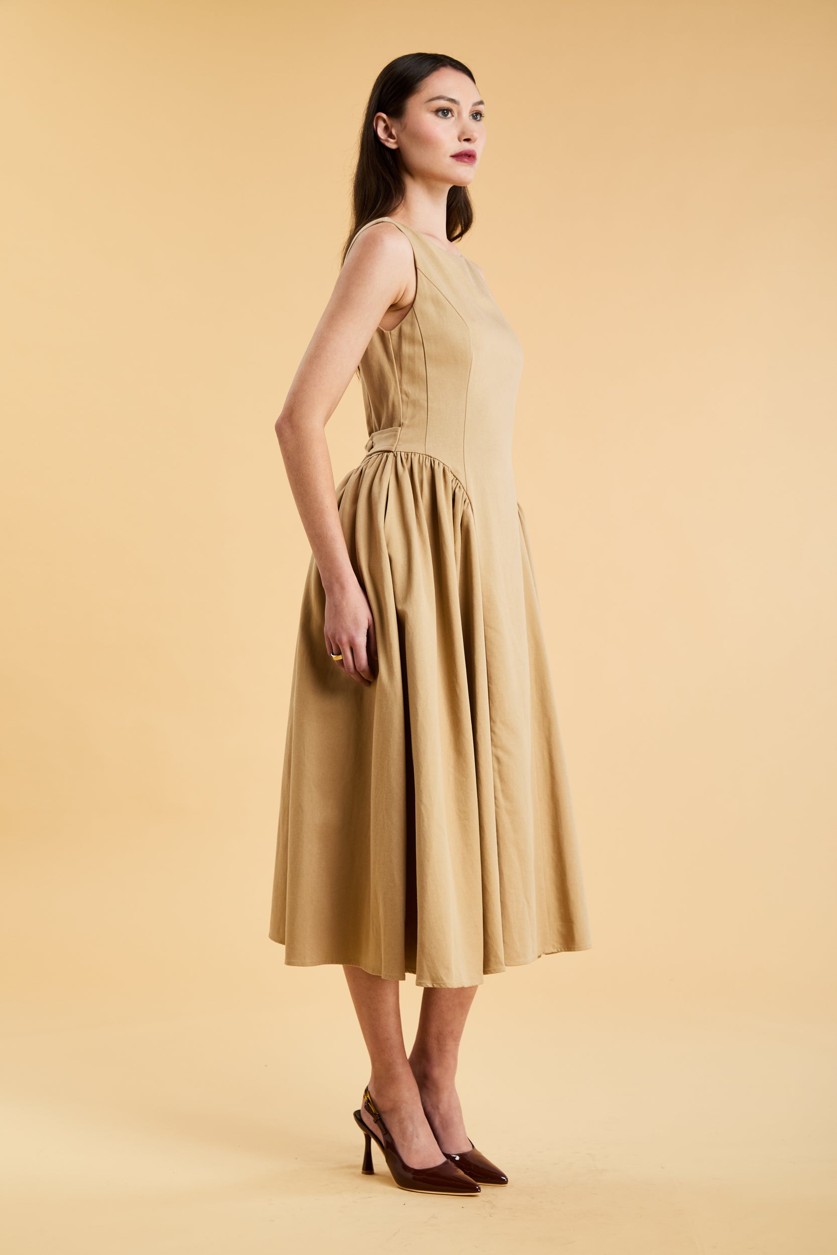 Woman wearing a Triplicity Camel colored sleeveless midi dress with gathered side panels on a beige background