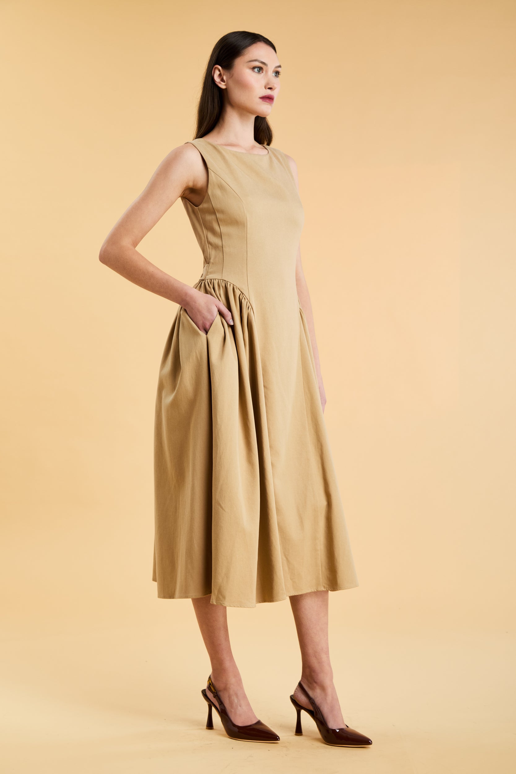 Woman wearing a Triplicity Camel colored sleeveless midi dress with gathered side panels on a beige background