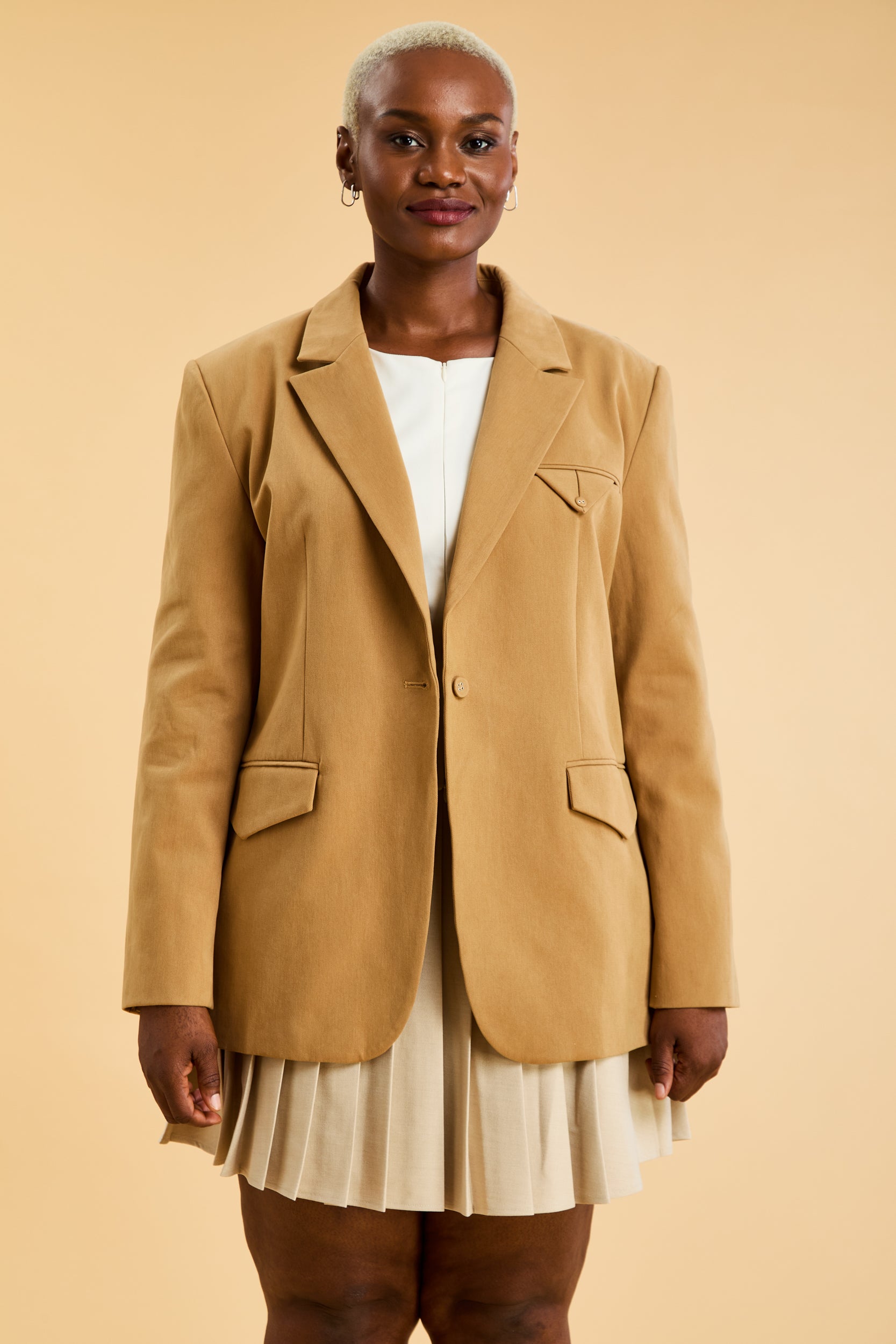 Woman wearing a Camel colored oversized blazer over a white top and pleated skirt on a beige background