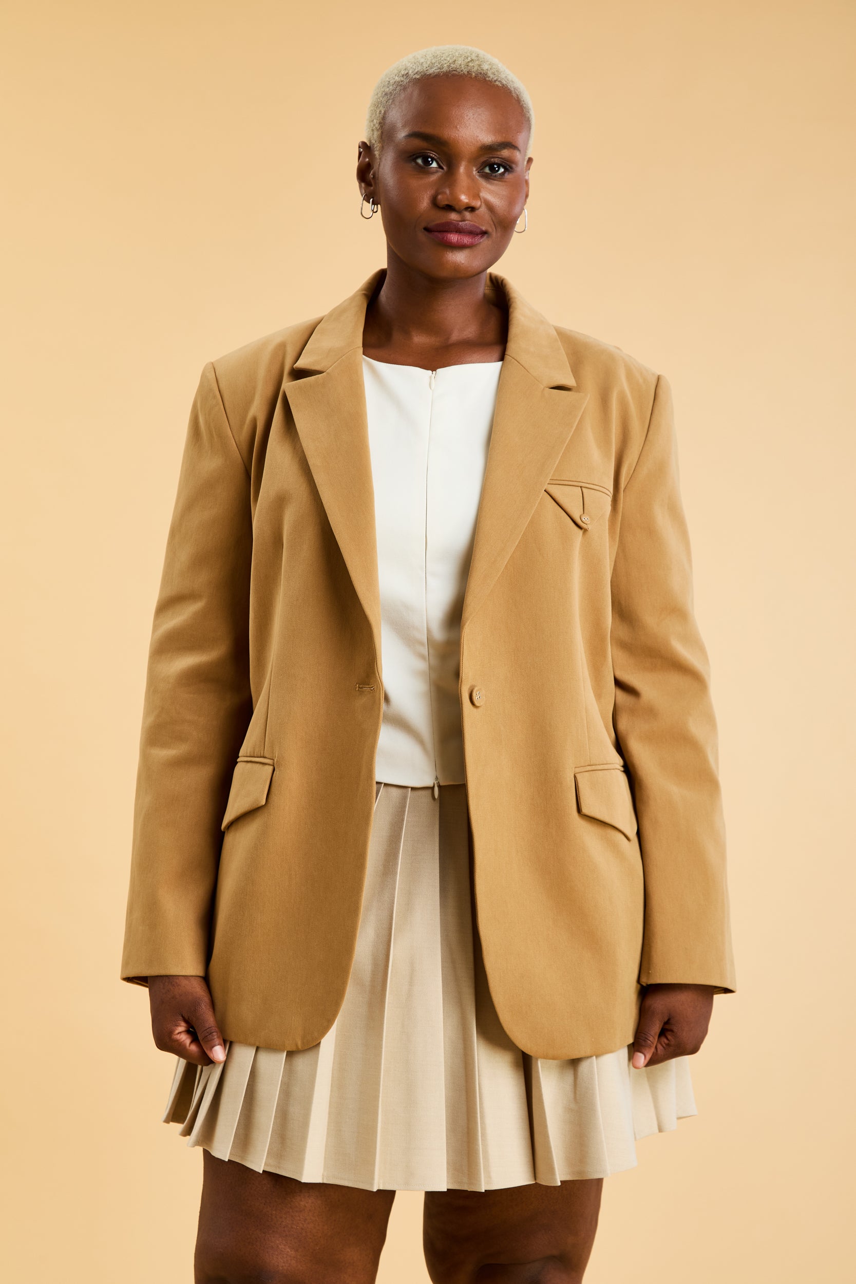 Woman wearing a beige blazer over a white top and pleated skirt on a beige background
