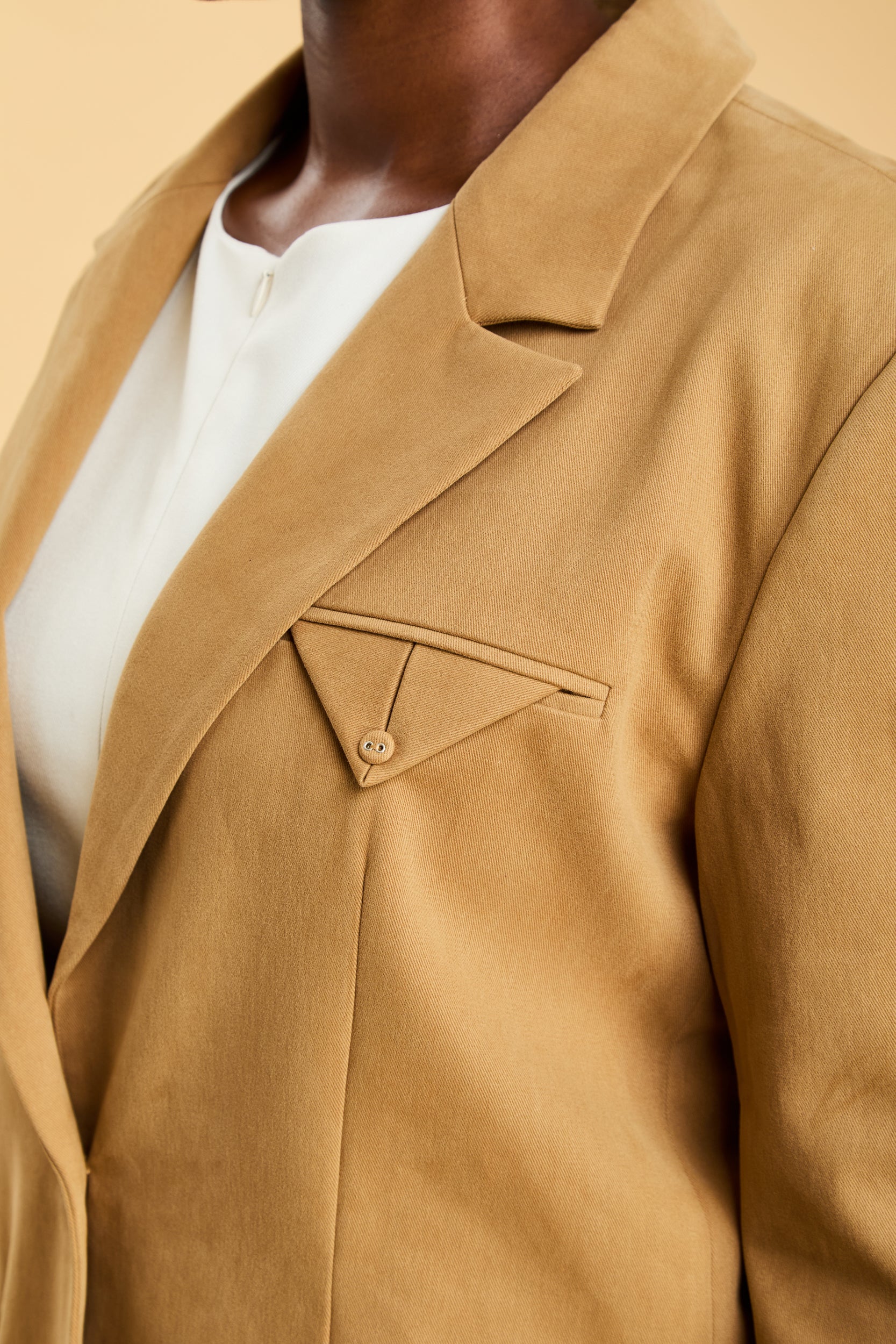 Close-up of a Camel colored blazer with a detailed welt pocket and button on a neutral background