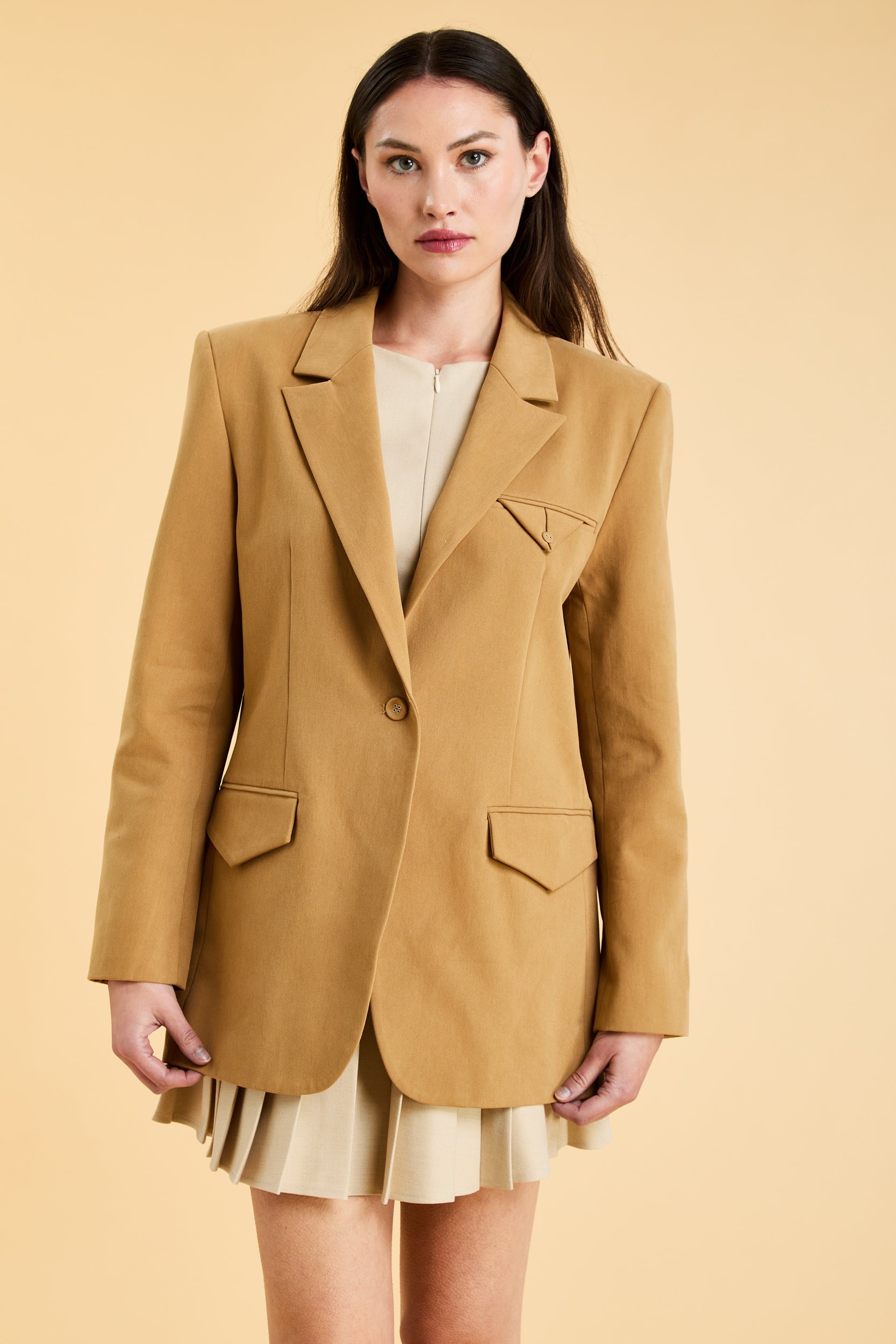 Woman wearing a tan Camel colored blazer over a beige top and skirt on a beige background