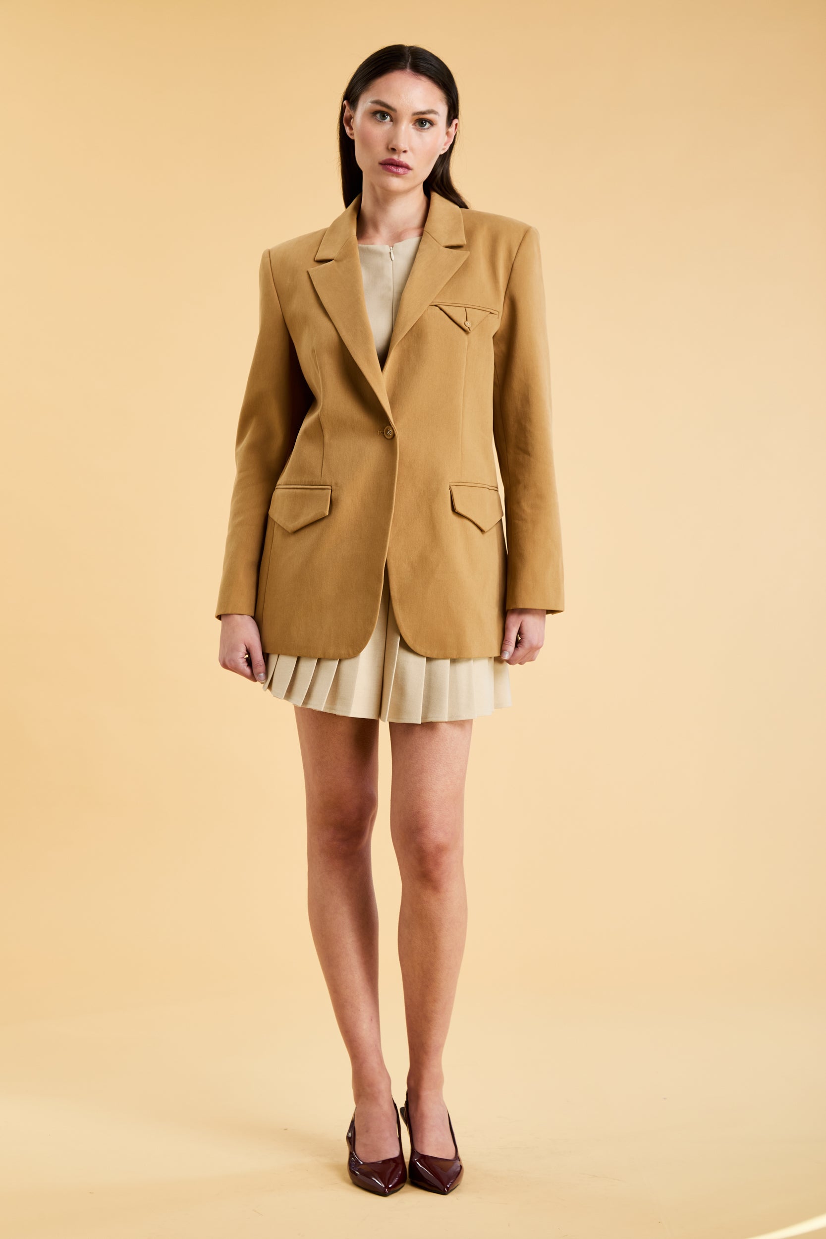 Woman wearing a Camel colored blazer over a white top and skirt on a beige background