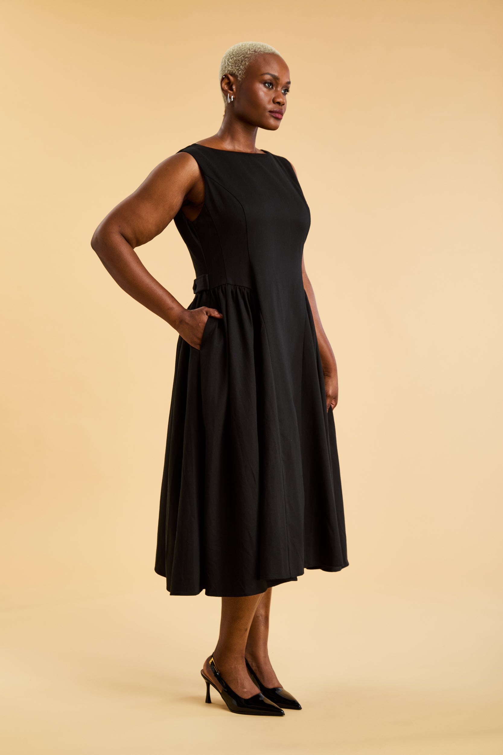 Woman wearing a Triplicity Black sleeveless midi dress with gathered side panels on a beige background