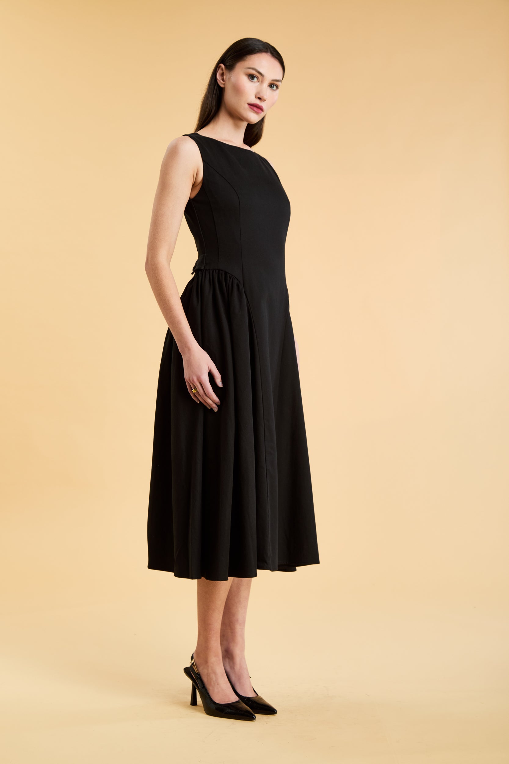 Woman wearing a Triplicity Black sleeveless midi dress with gathered side panels on a beige background