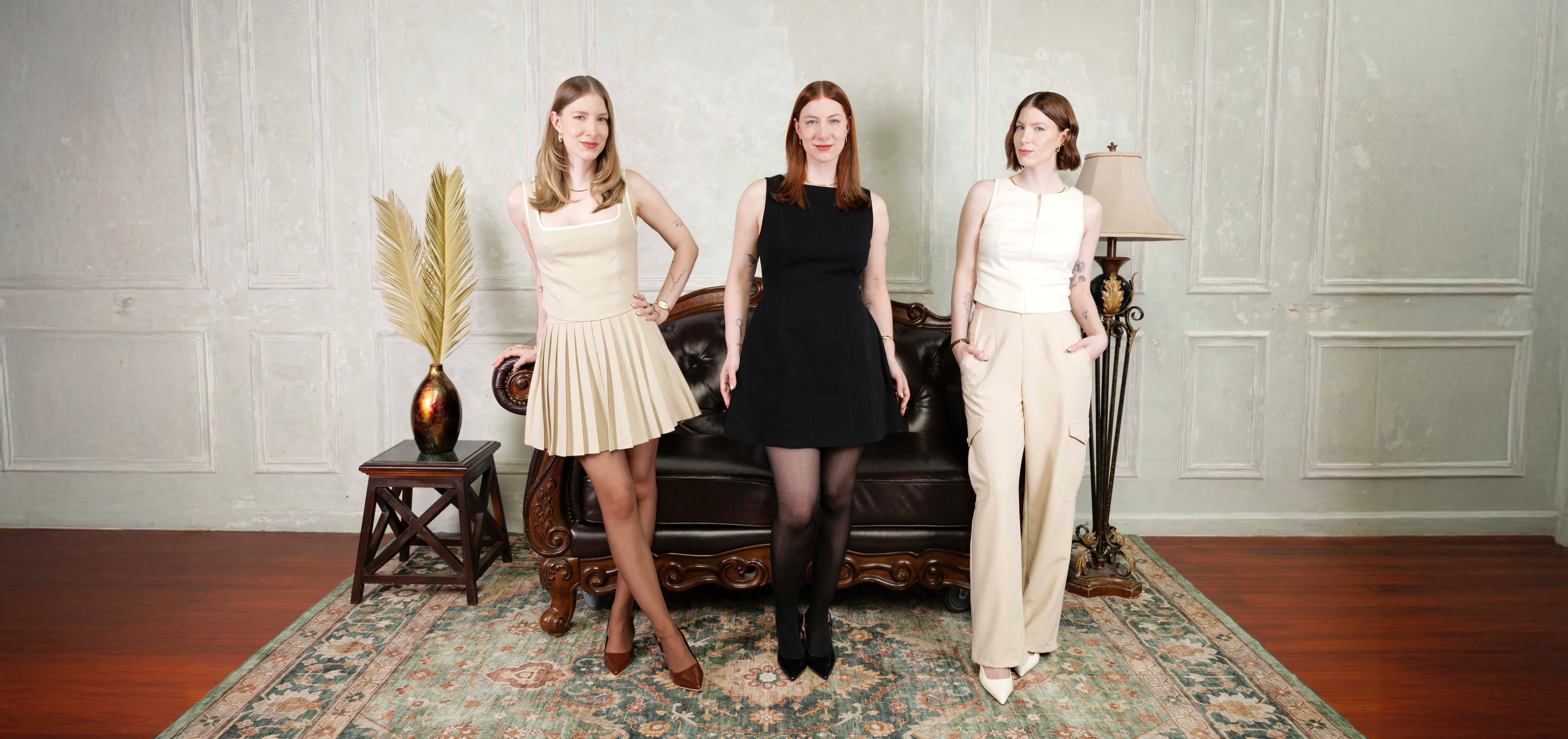 Three women (The Levesque Triplets) in formal attire (Triplicity Clothing) standing in a room with a sofa and decorative items.