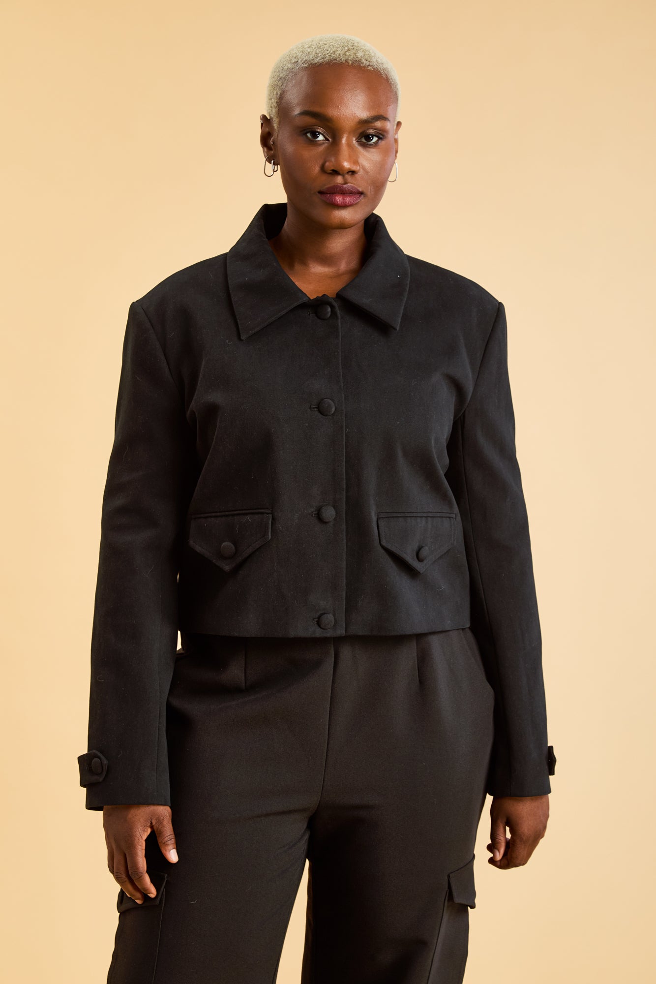 Woman wearing a Black Triplicity collared jacket and Black Triplicity pants on a beige background front close up