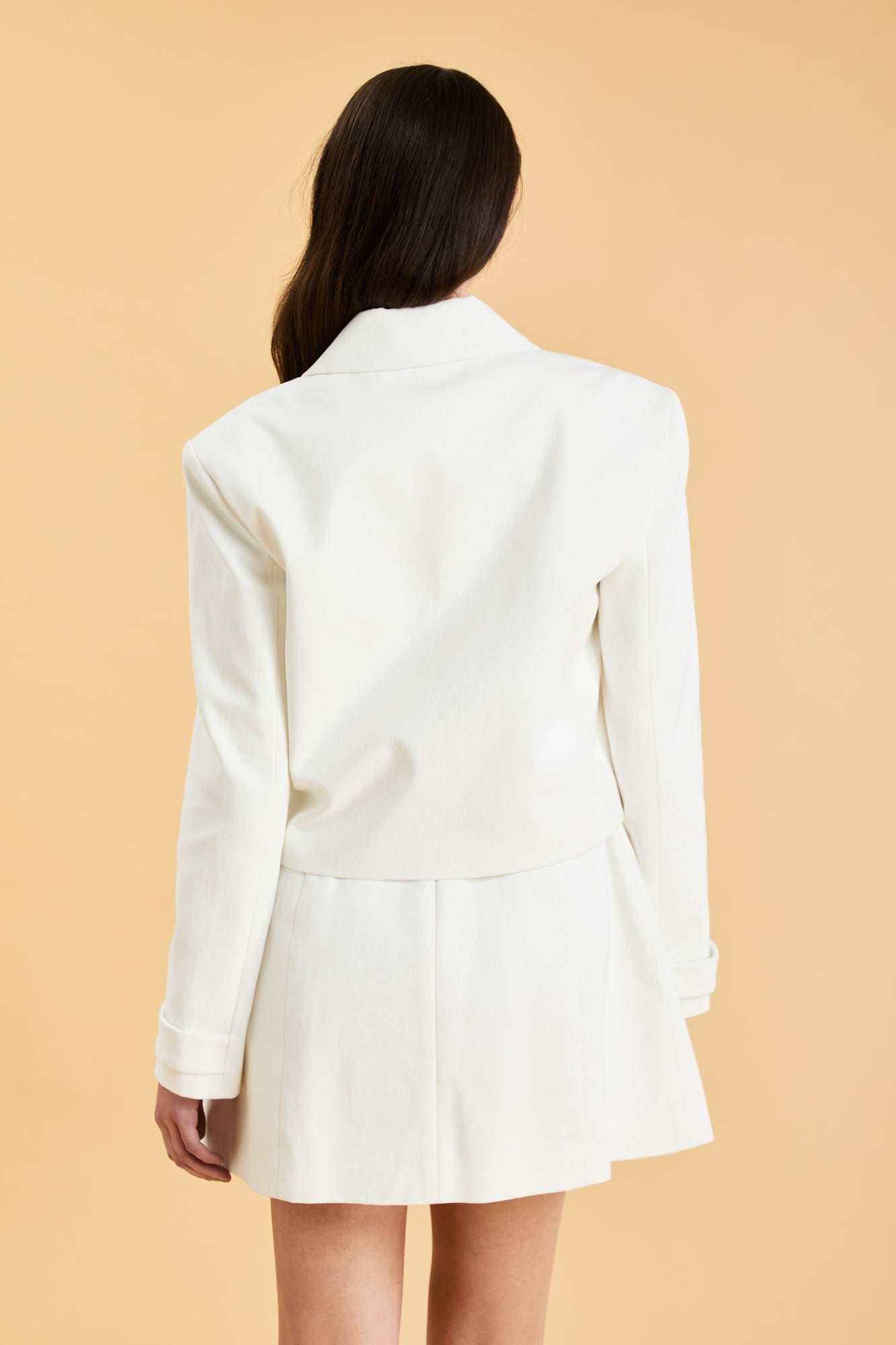 Woman wearing a White Triplicity collared tailored jacket against a beige background back view close up
