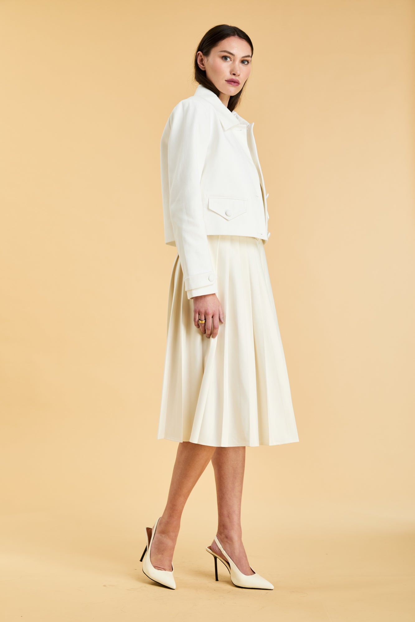 Woman wearing a White Triplicity collared tailored jacket and a White Triplicity pleated skirt against a beige background side view full body