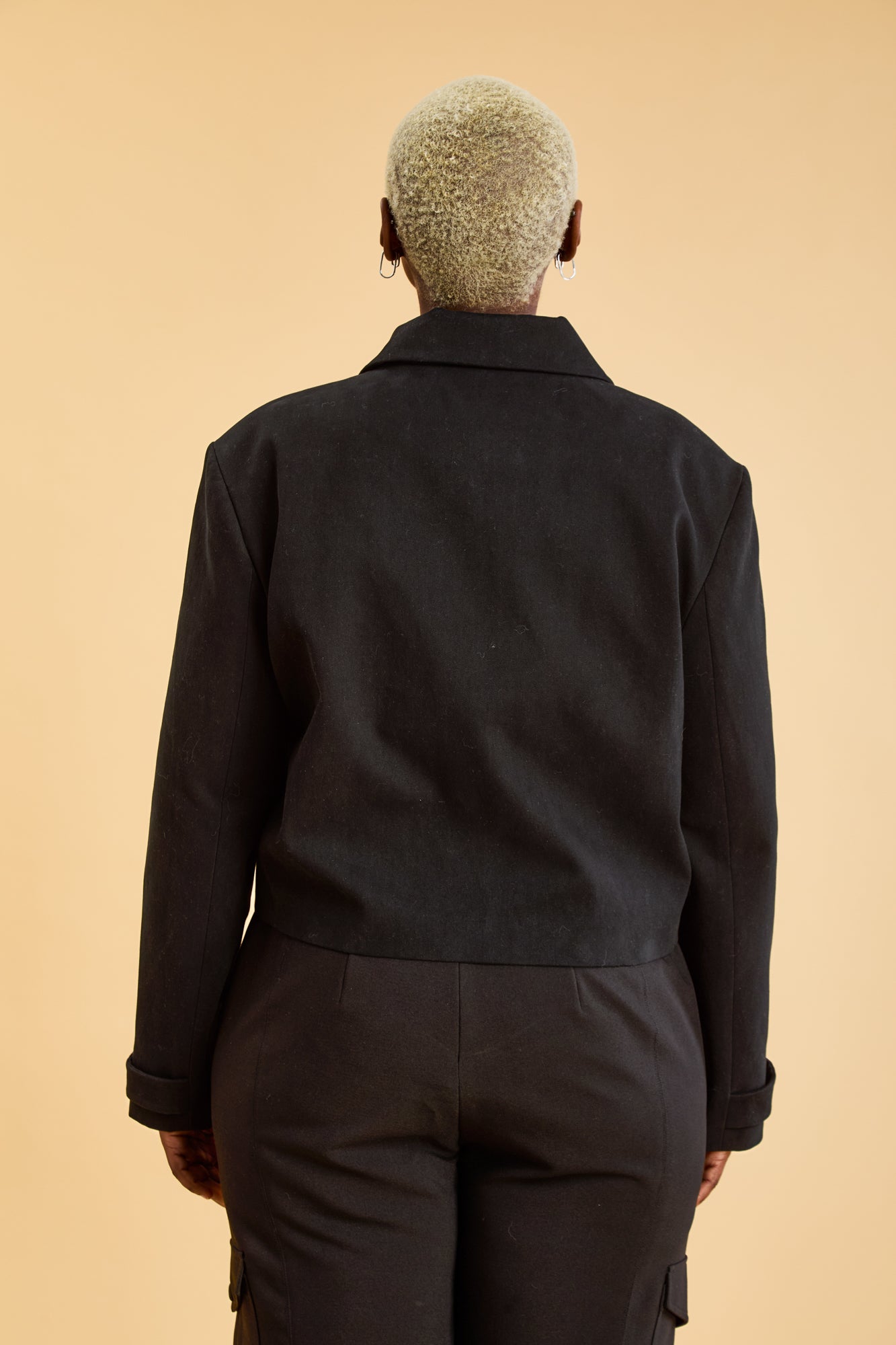 Woman wearing a Black Triplicity collared jacket and Black Triplicity pants on a beige background back close up