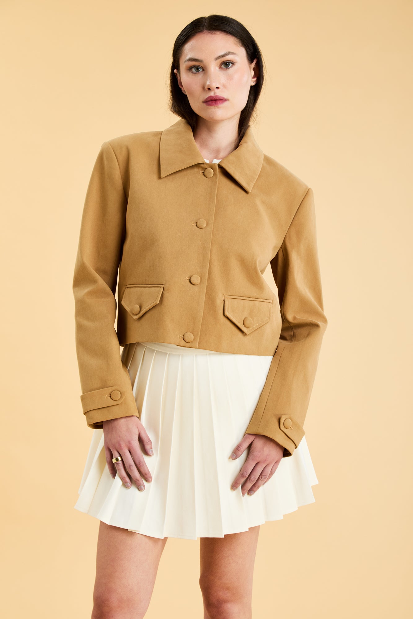 Woman wearing a Camel colored Triplicity collared tailored jacket on top of a White Triplicity pleated mini skirt on a beige background front view close up