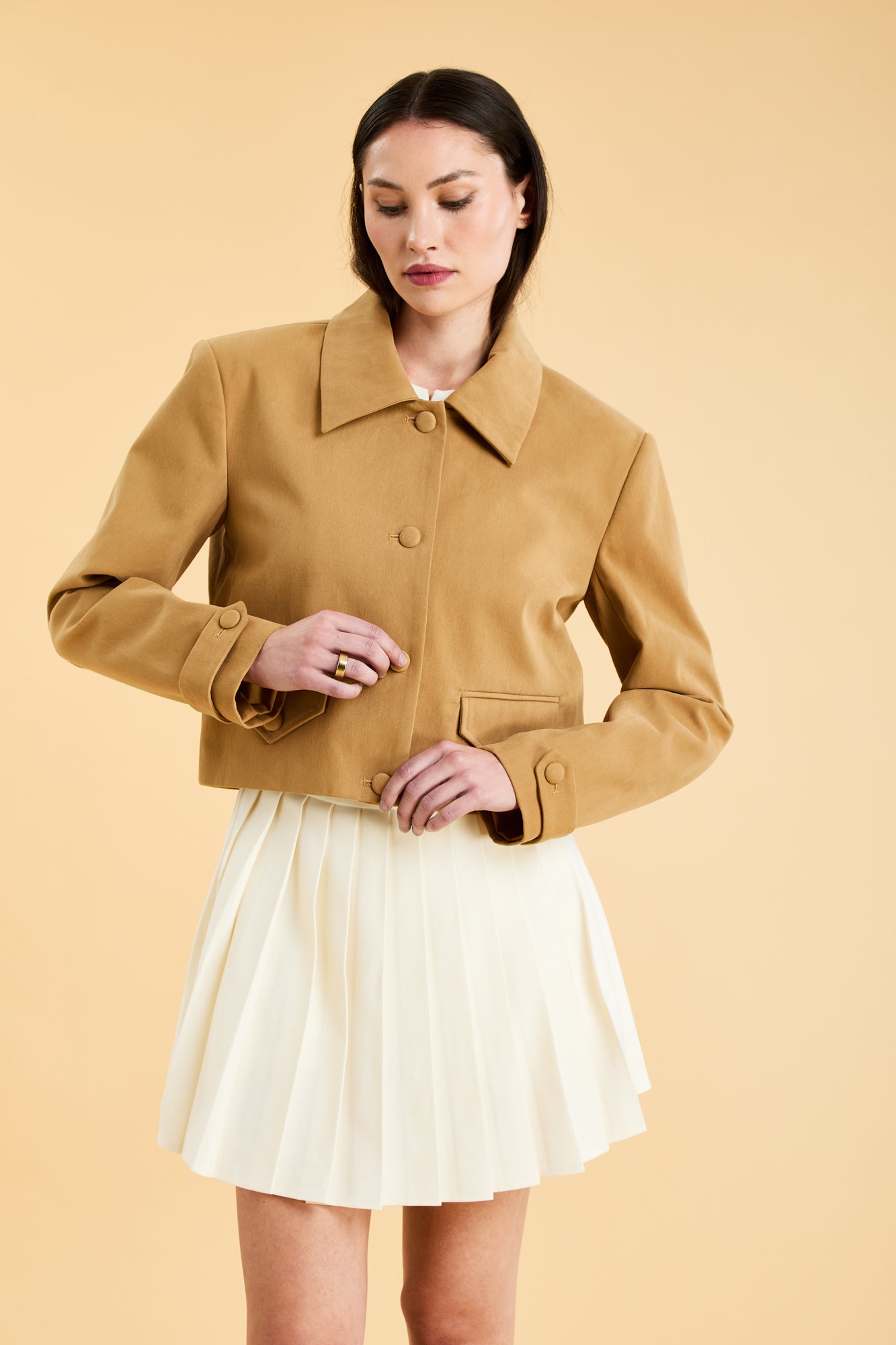 Woman wearing a Camel colored Triplicity collared tailored jacket on top of a White Triplicity pleated mini skirt on a beige background front view close up