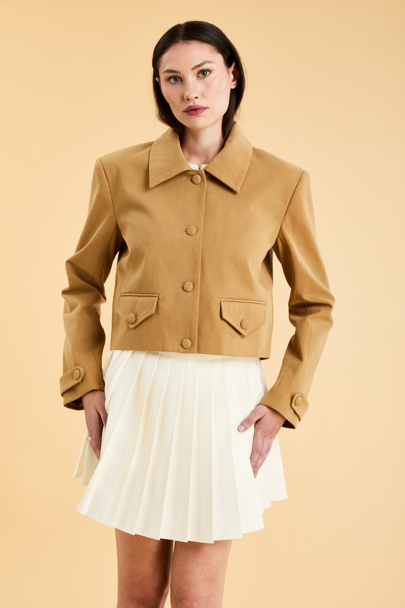 Woman wearing a Camel colored Triplicity collared tailored jacket on top of a White Triplicity pleated mini skirt on a beige background front view close up