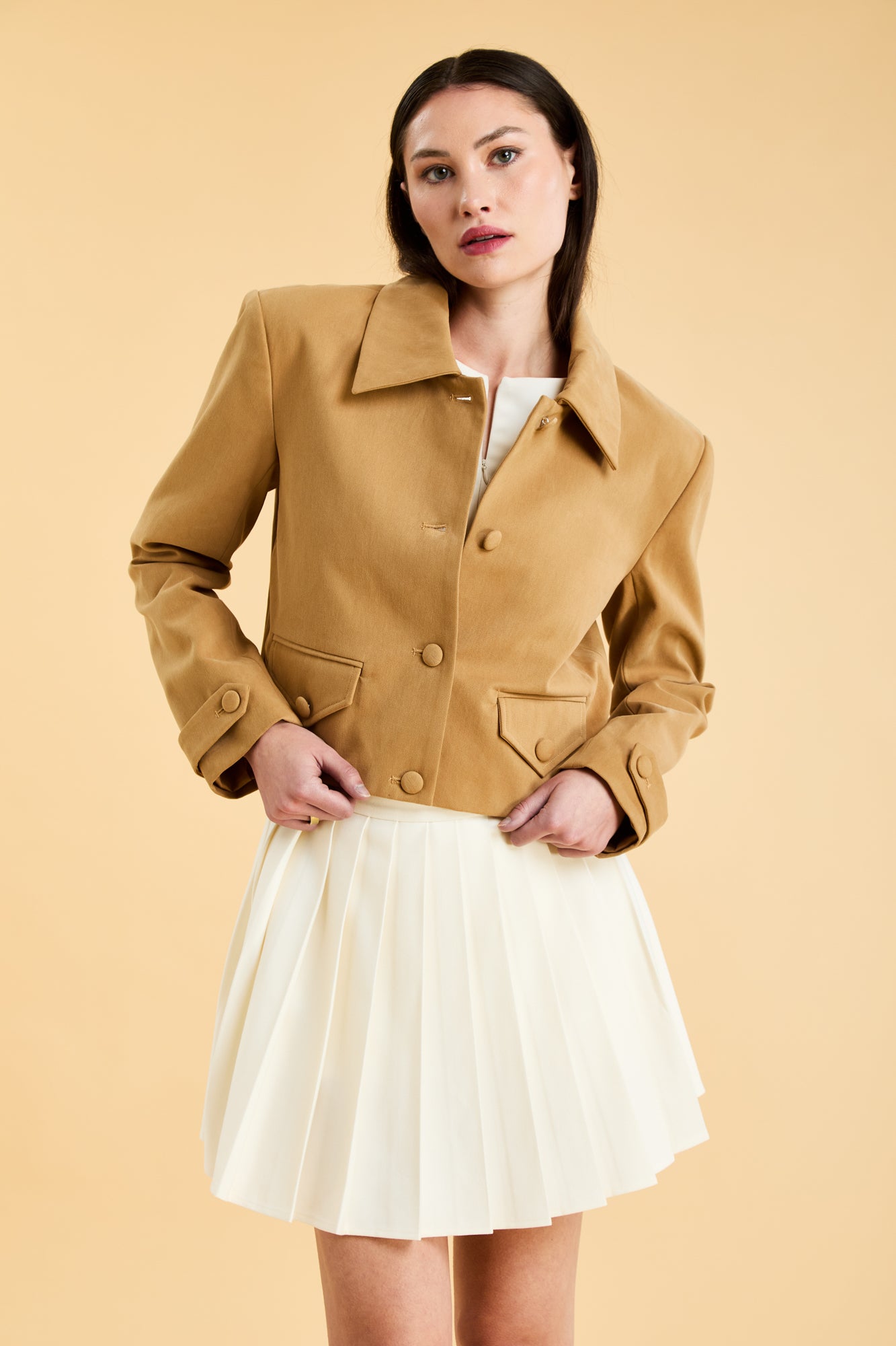Woman wearing a Camel colored Triplicity collared tailored jacket on top of a White Triplicity pleated mini skirt on a beige background front view close up