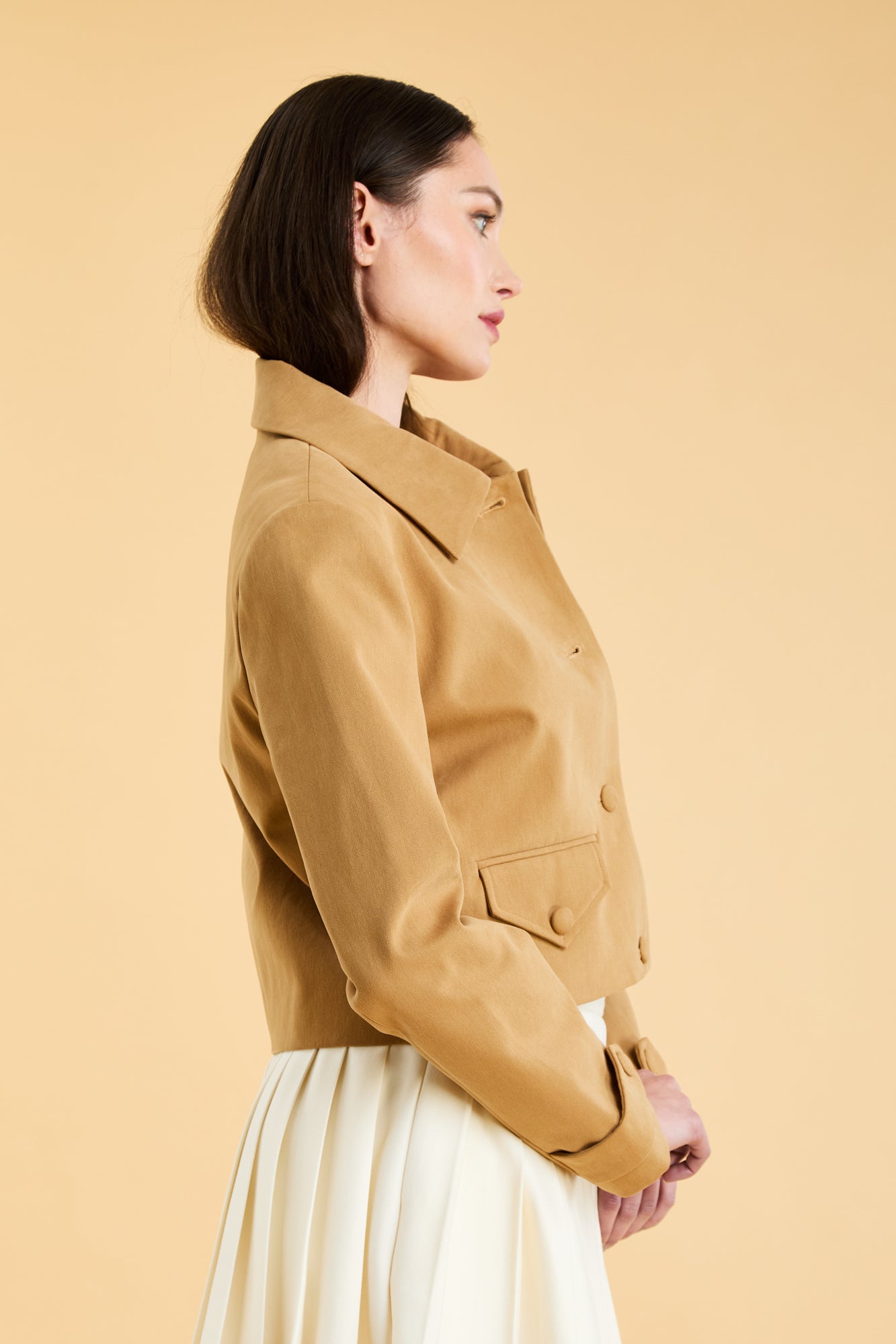 Woman wearing a Camel colored Triplicity collared tailored jacket on top of a White Triplicity pleated mini skirt on a beige background side view close up