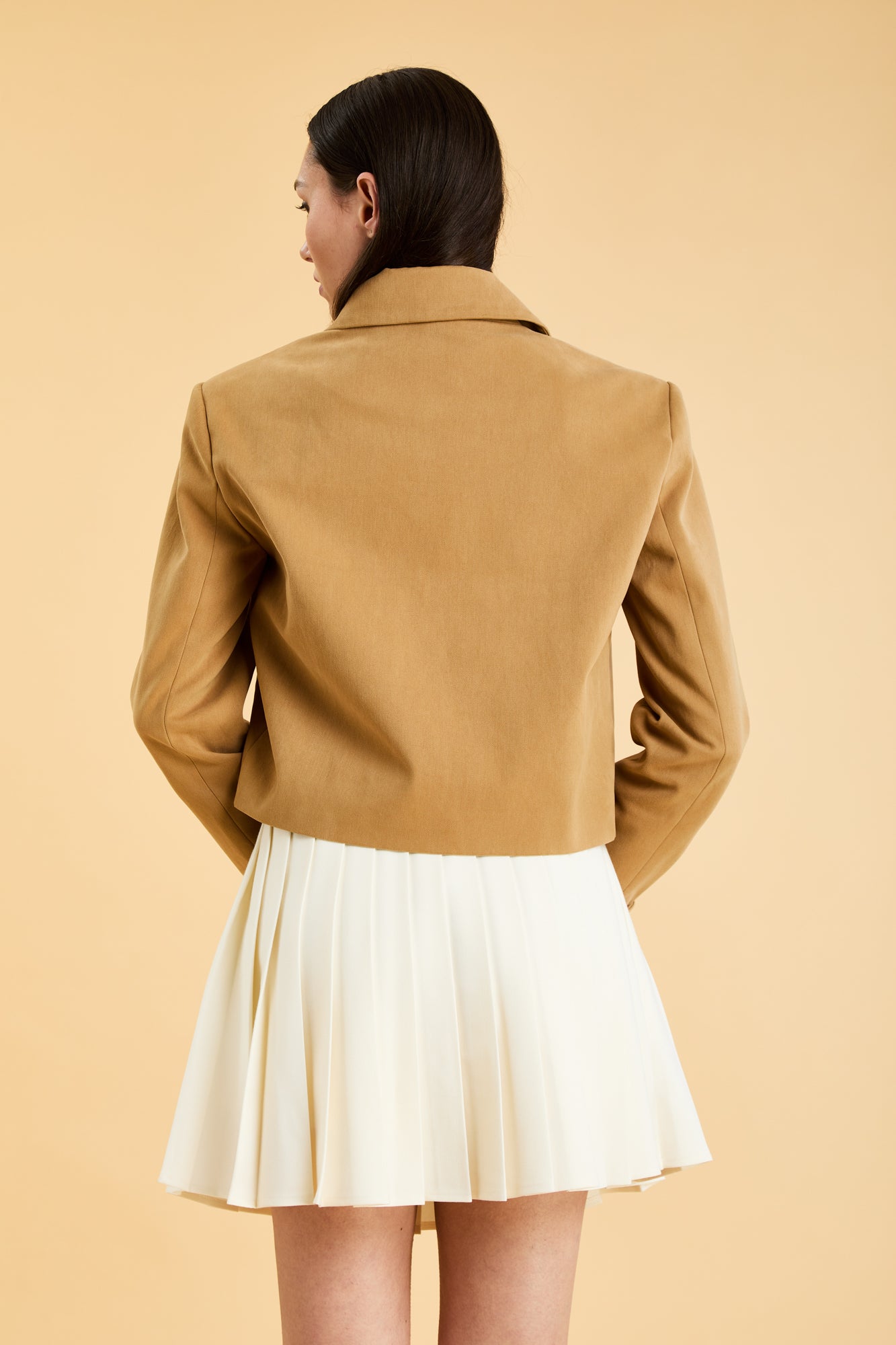 Woman wearing a Camel colored Triplicity collared tailored jacket on top of a White Triplicity pleated mini skirt on a beige background back view close up