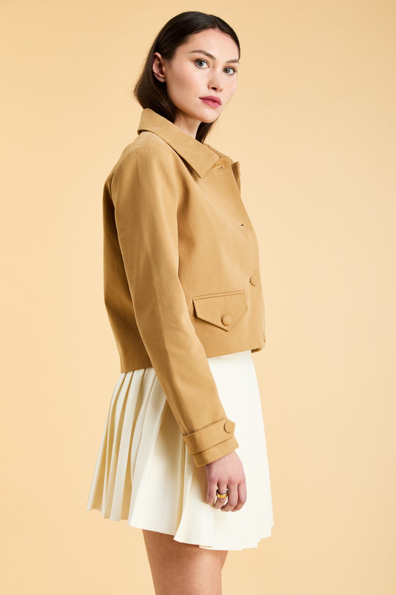 Woman wearing a Camel colored Triplicity collared tailored jacket on top of a White Triplicity pleated mini skirt on a beige background side view close up
