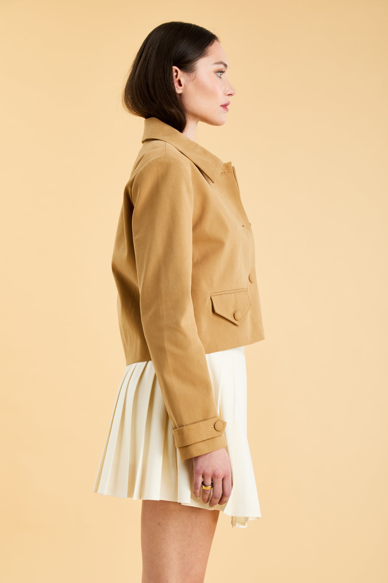 Woman wearing a Camel colored Triplicity collared tailored jacket on top of a White Triplicity pleated mini skirt on a beige background side view close up