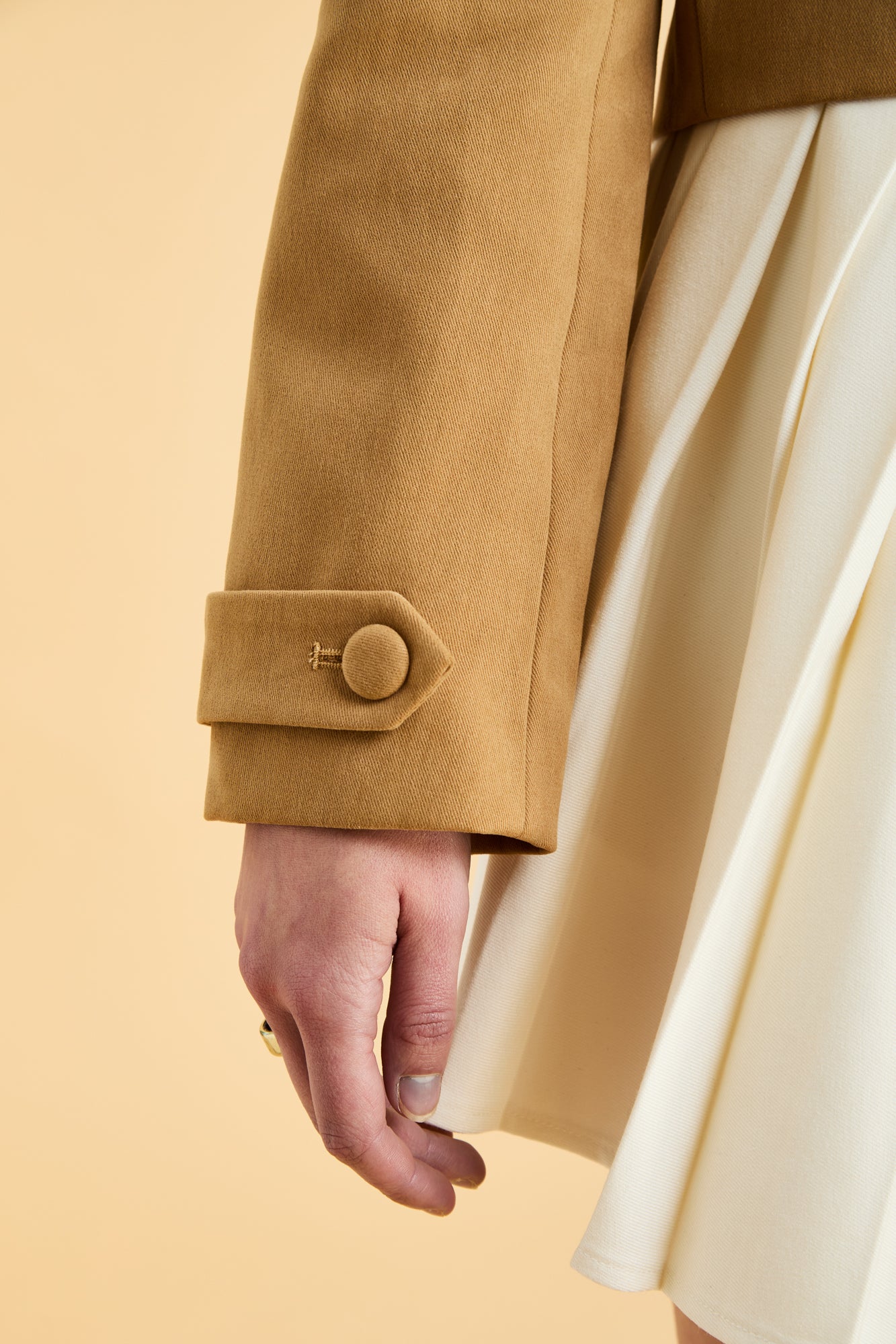 Close-up of a person wearing a Camel colored jacket with a button, holding a beige fabric against a beige background
