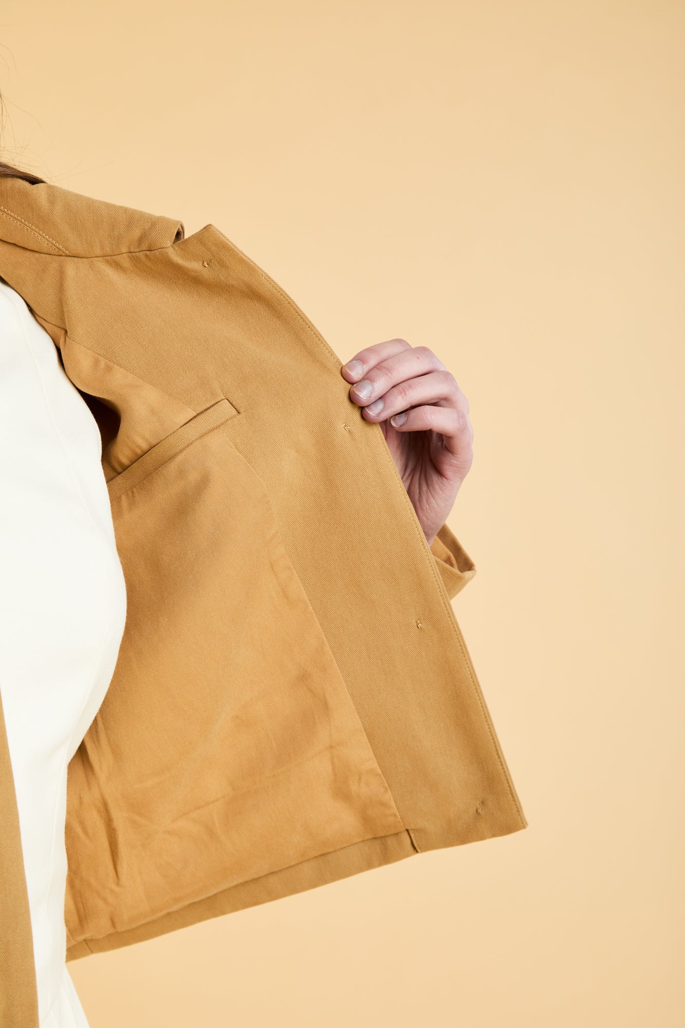 Camel colored Triplicity jacket with a hand holding the jacket edge and showing the interior pocket on a beige background