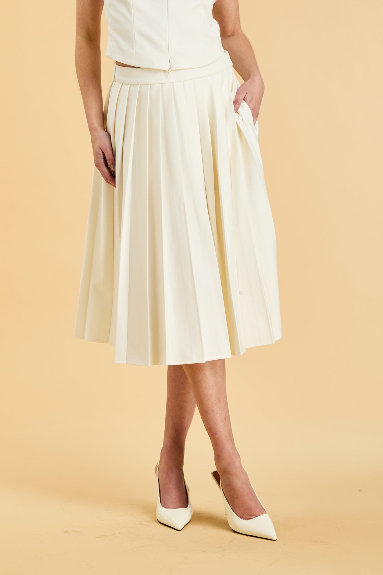 Close up of the bottom half of a woman wearing a White colored Triplicity Alice top and White Triplicity pleated midi skirt against a beige background front view