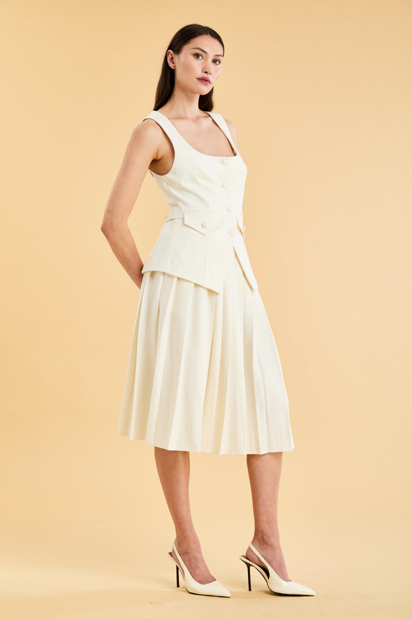 Woman wearing a White colored Triplicity Georgiana vest top and White Triplicity pleated midi skirt against a beige background three quarter view full body