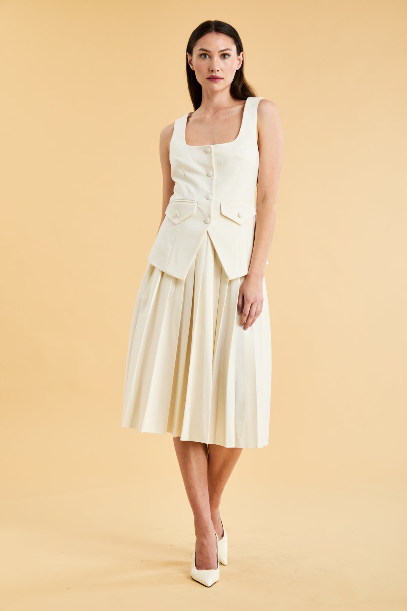 Woman wearing a White colored Triplicity Georgiana vest top and White Triplicity pleated midi skirt against a beige background front view full body