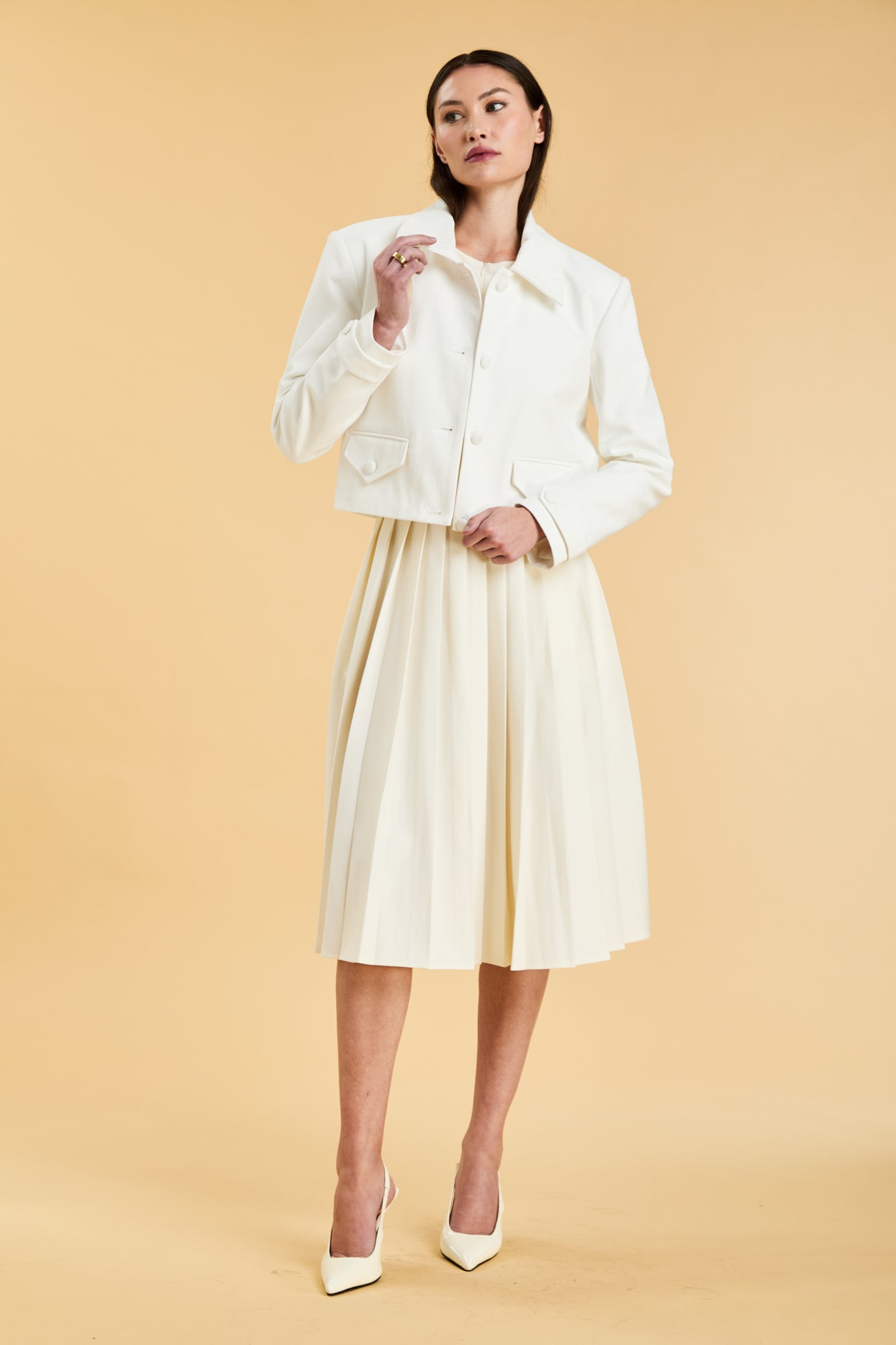 Woman wearing a White colored Triplicity Charlotte collared cropped jacket and White Triplicity pleated midi skirt with slit against a beige background front view full body