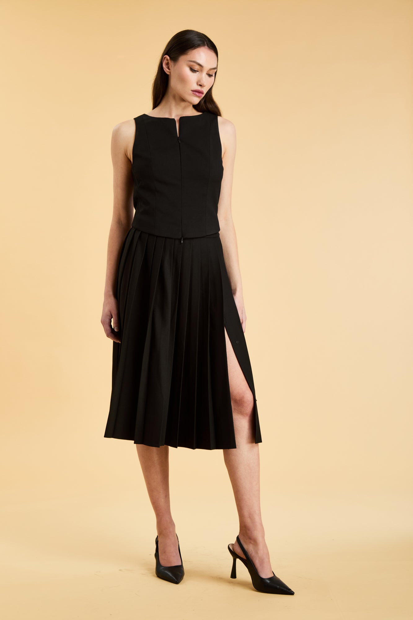 Woman wearing a black sleeveless top on a pleated Black midi skirt on a beige background
