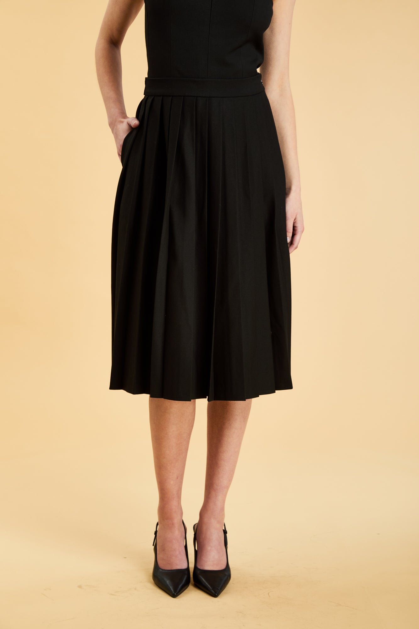 Woman wearing a black sleeveless top on a pleated Black midi skirt on a beige background
