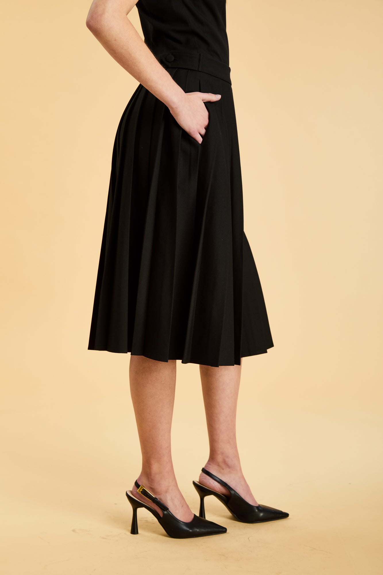 Woman wearing a black sleeveless top on a pleated Black midi skirt on a beige background