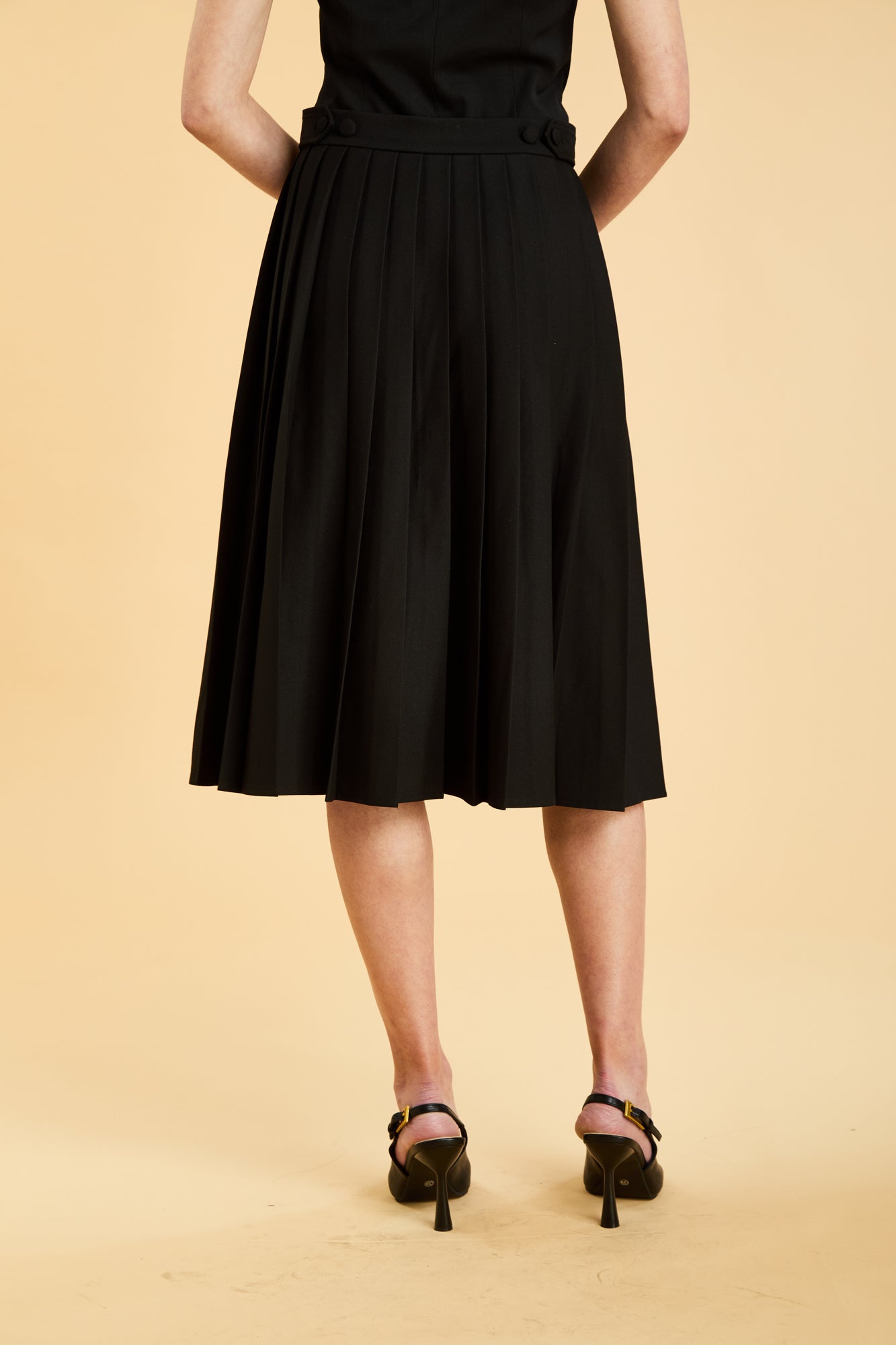 Woman wearing a black sleeveless top on a pleated Black midi skirt on a beige background