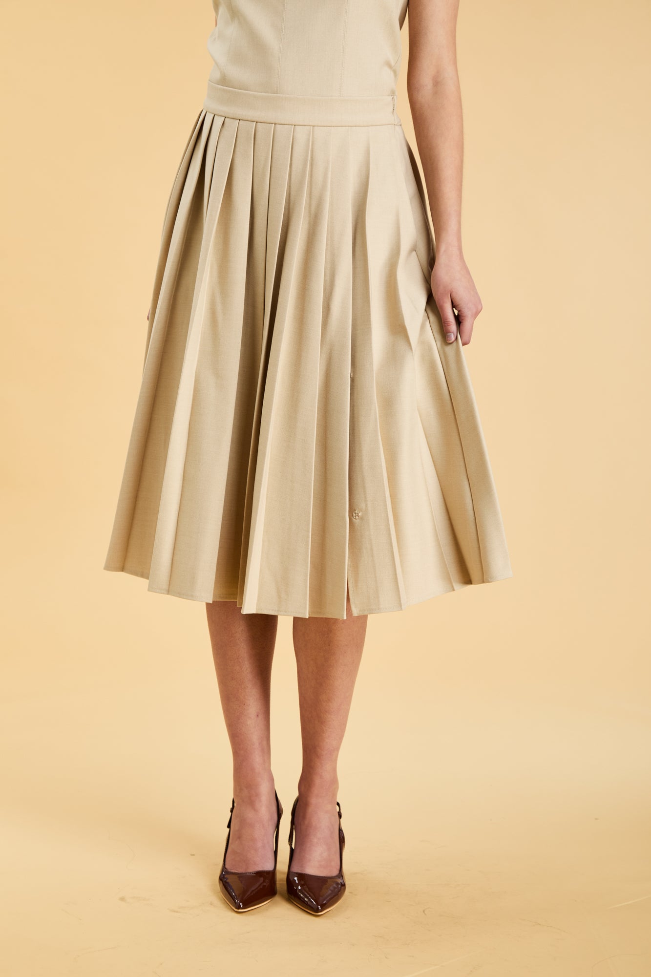 Close up of the bottom half of a woman wearing a Triplicity Oat Elisabeth pleated mini skirt against a beige background front view