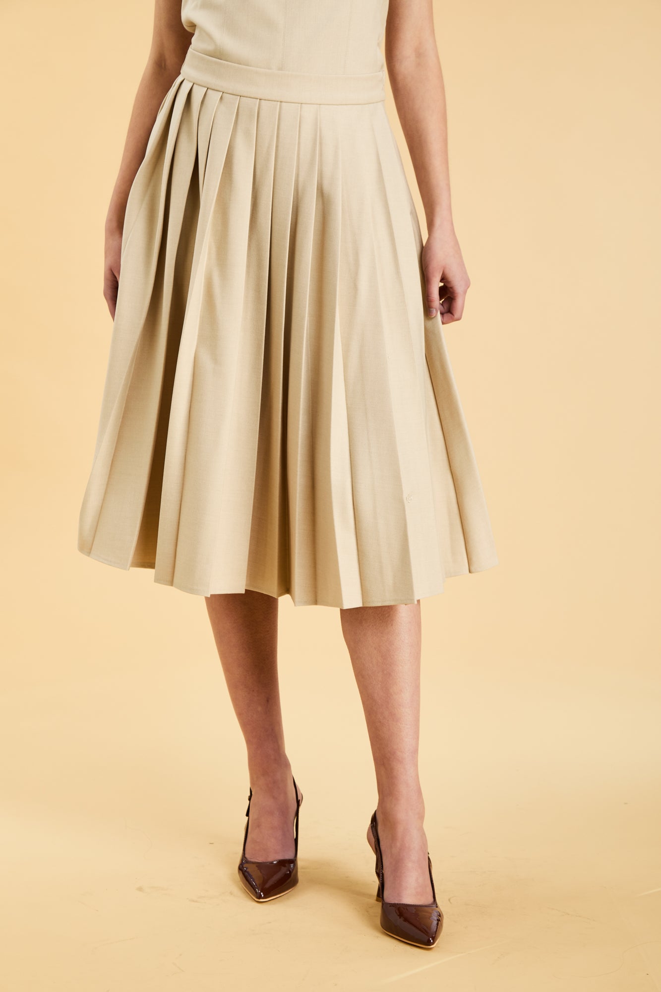 Close up of the bottom half of a woman wearing a Triplicity Oat Elisabeth pleated mini skirt against a beige background front view