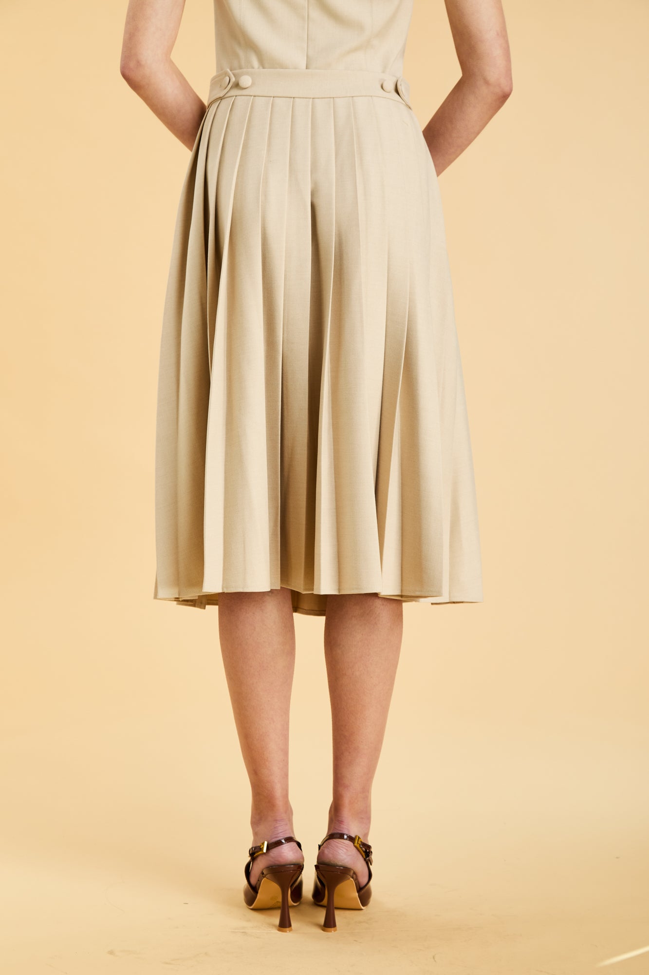 Close up of the bottom half of a woman wearing a Triplicity Oat Elisabeth pleated mini skirt against a beige background back view