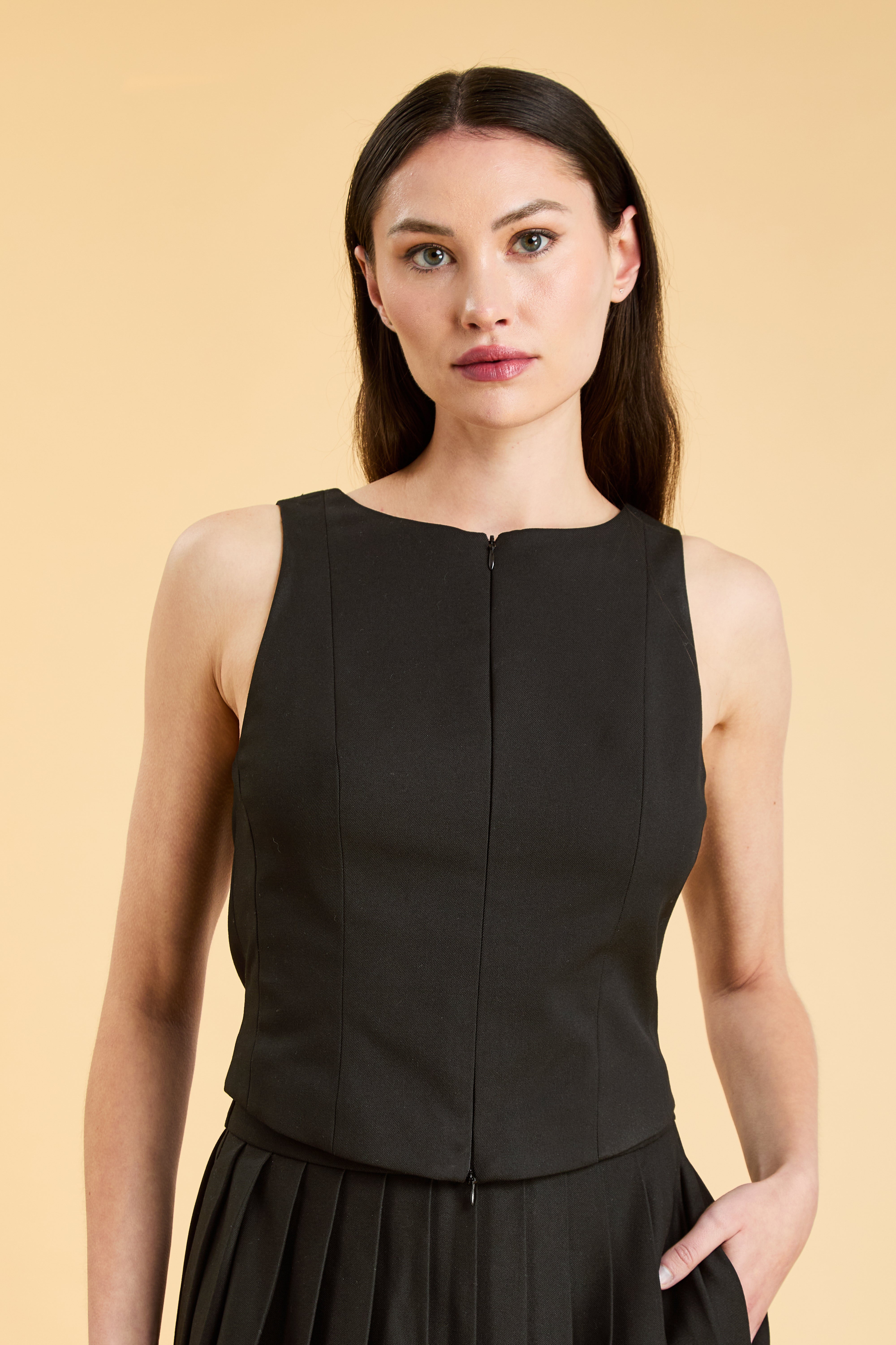 Person wearing a Black Alice double ended zipper Top on a Black pleated Elisabeth Mini skirt against a beige background