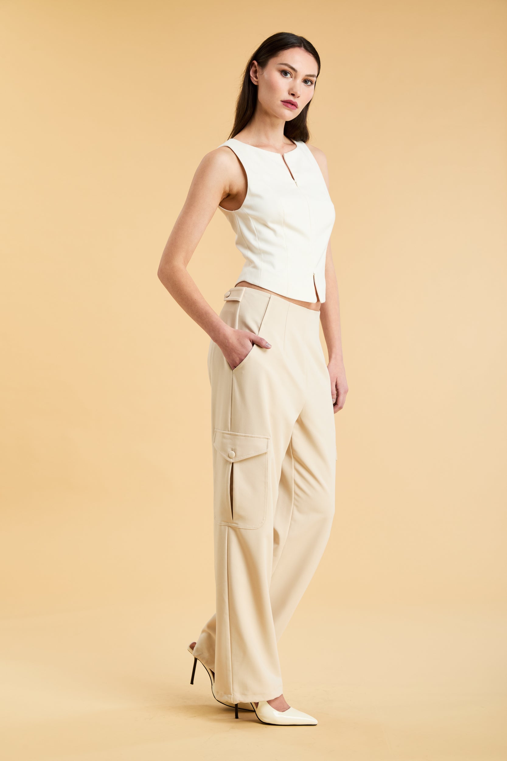 Woman wearing a white sleeveless double zipper top and beige almond Triplicity trousers cargo pants on a beige background