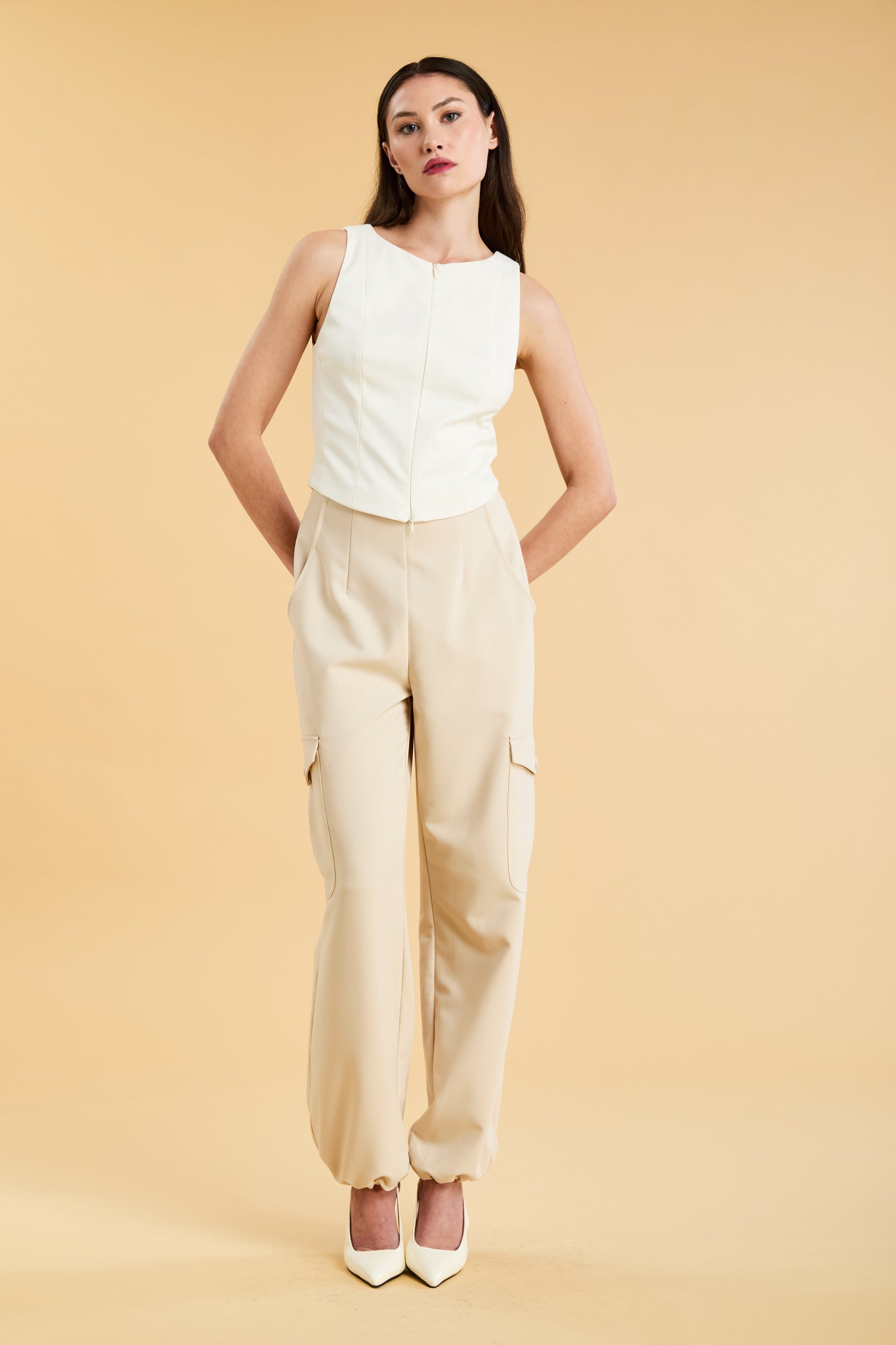 Woman wearing a white sleeveless double zipper top and beige almond Triplicity trousers cargo pants on a beige background