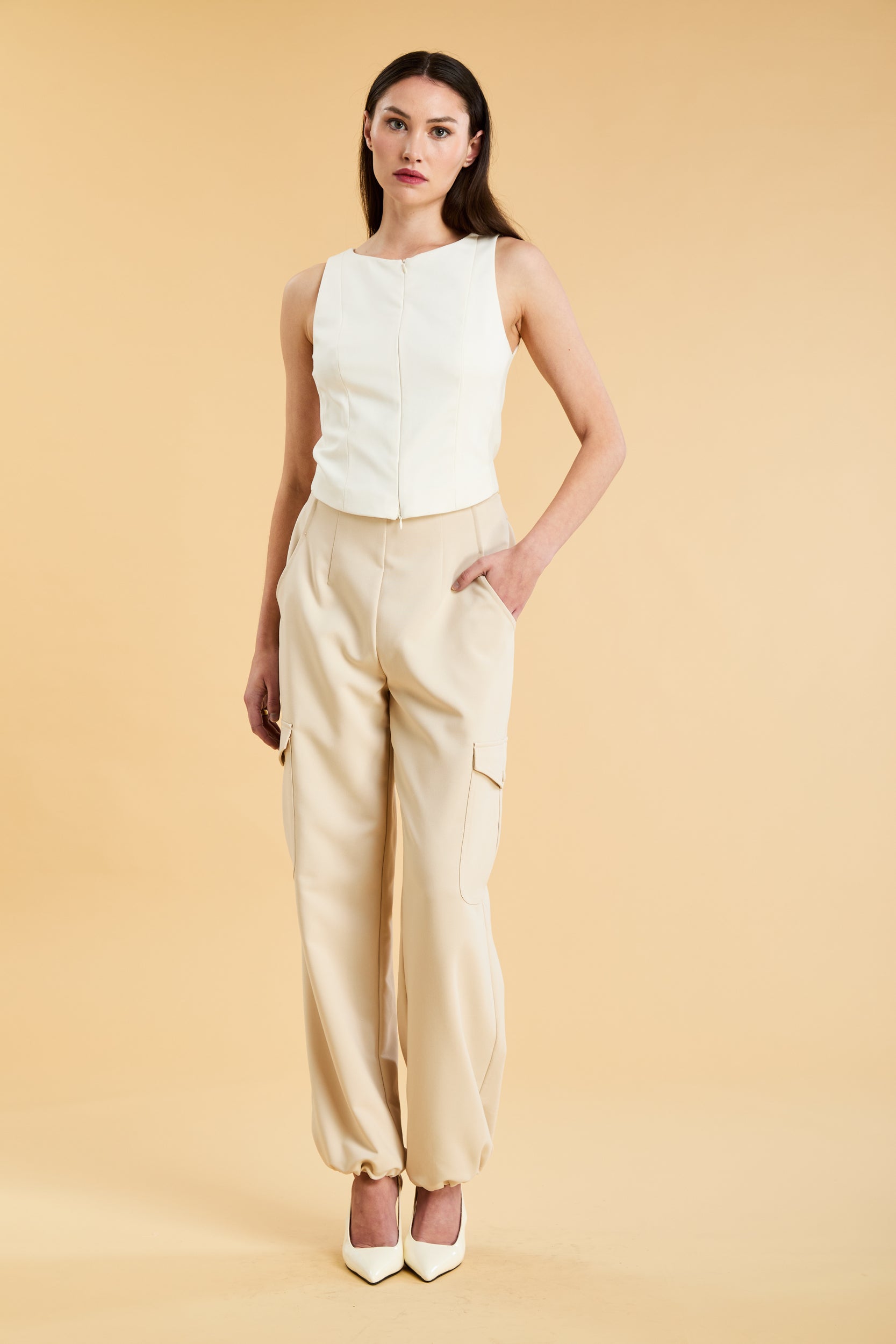 Woman wearing a white sleeveless double zipper top and beige almond Triplicity trousers cargo pants on a beige background